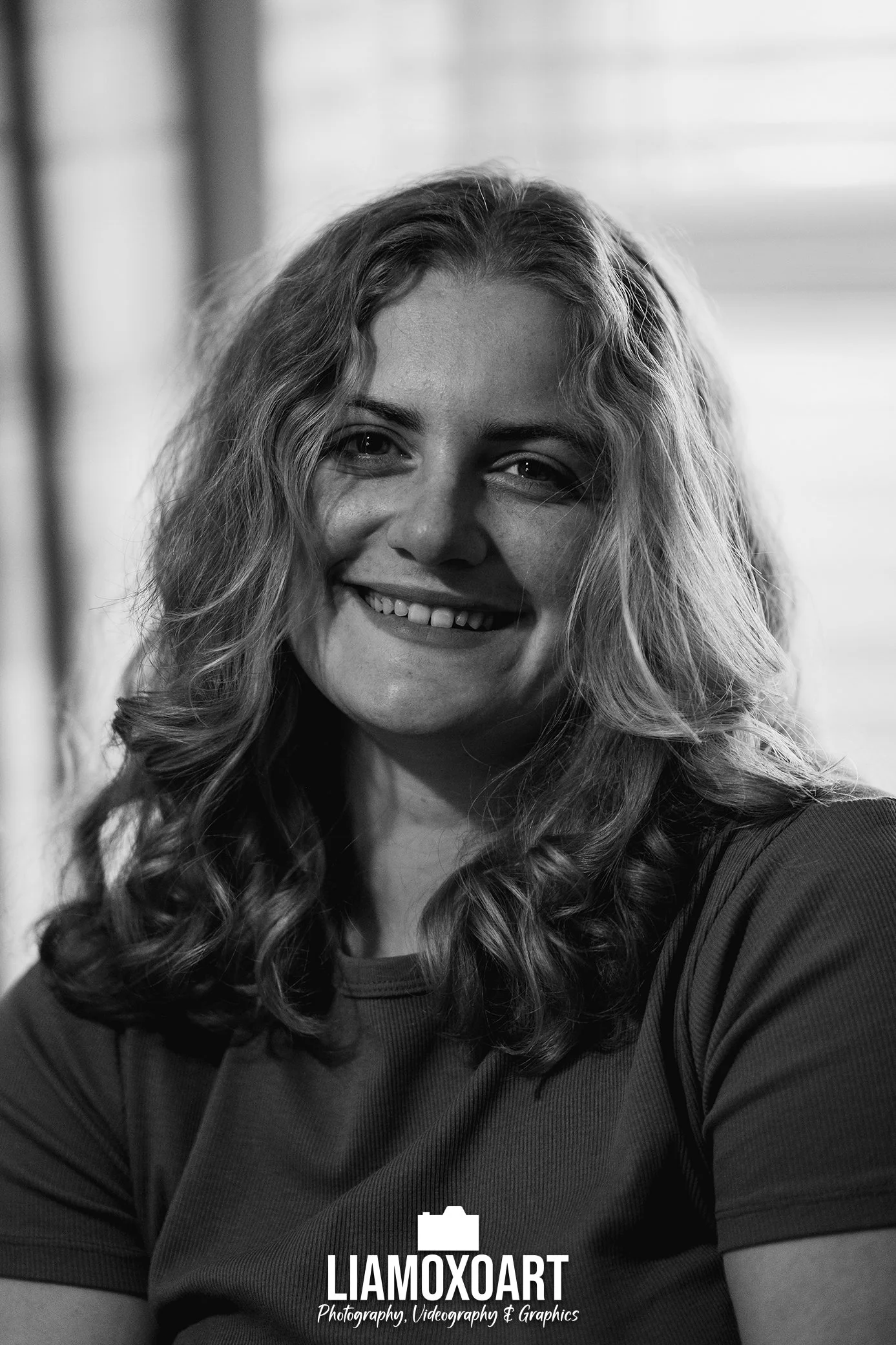 Black and white portrait of a smiling woman with long, curly hair, wearing a dark shirt, with a blurred background. The photograph is centered on her face and upper torso.