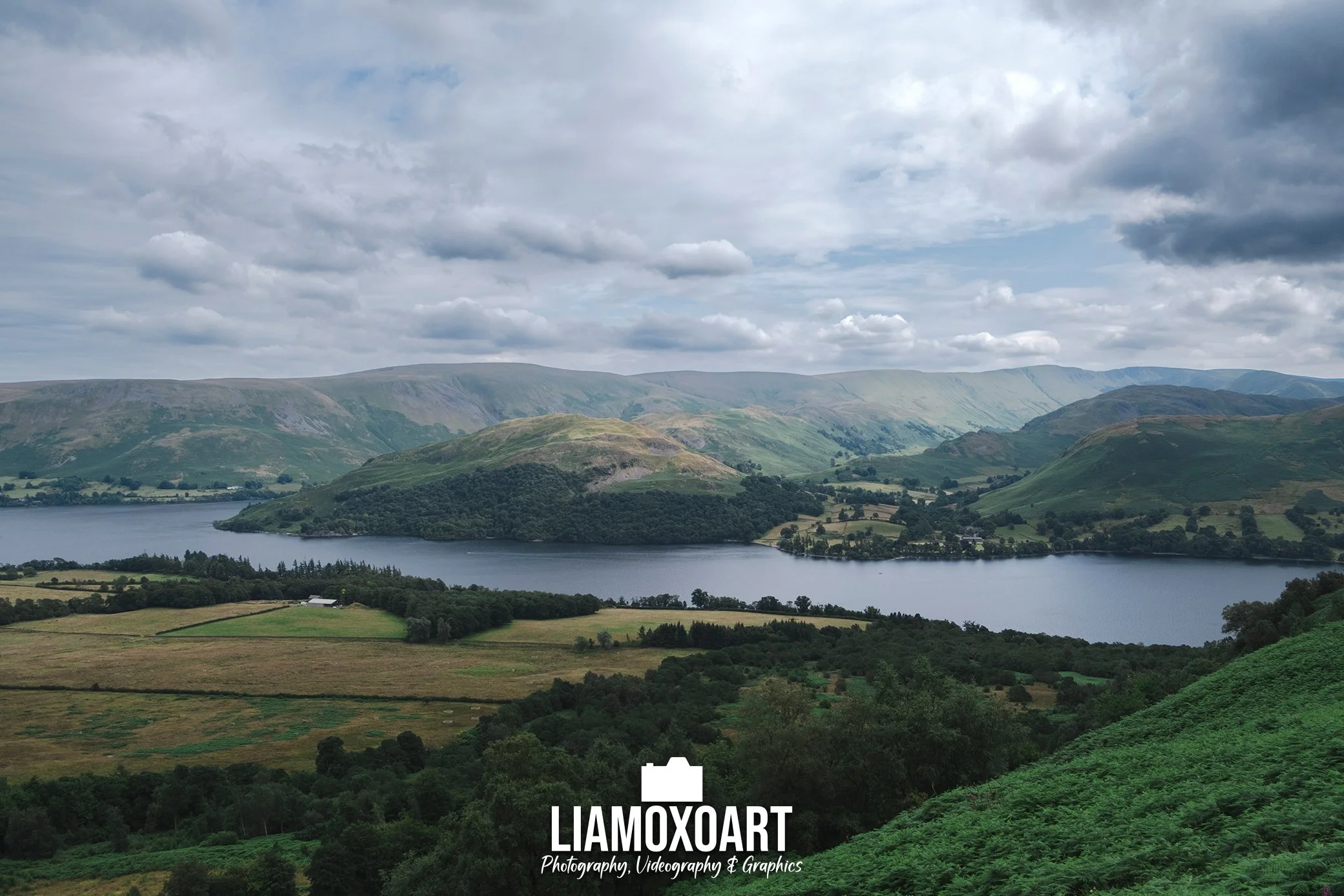 Scenic landscape of rolling green hills and a lake under a cloudy sky.
