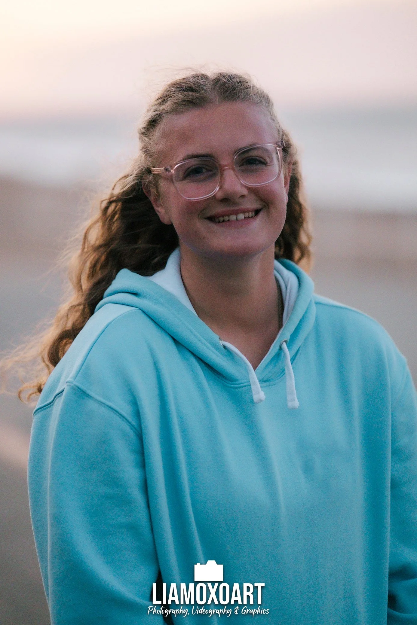 A young woman with curly hair, glasses, and a bright smile wearing a light blue hoodie outdoors during sunset.