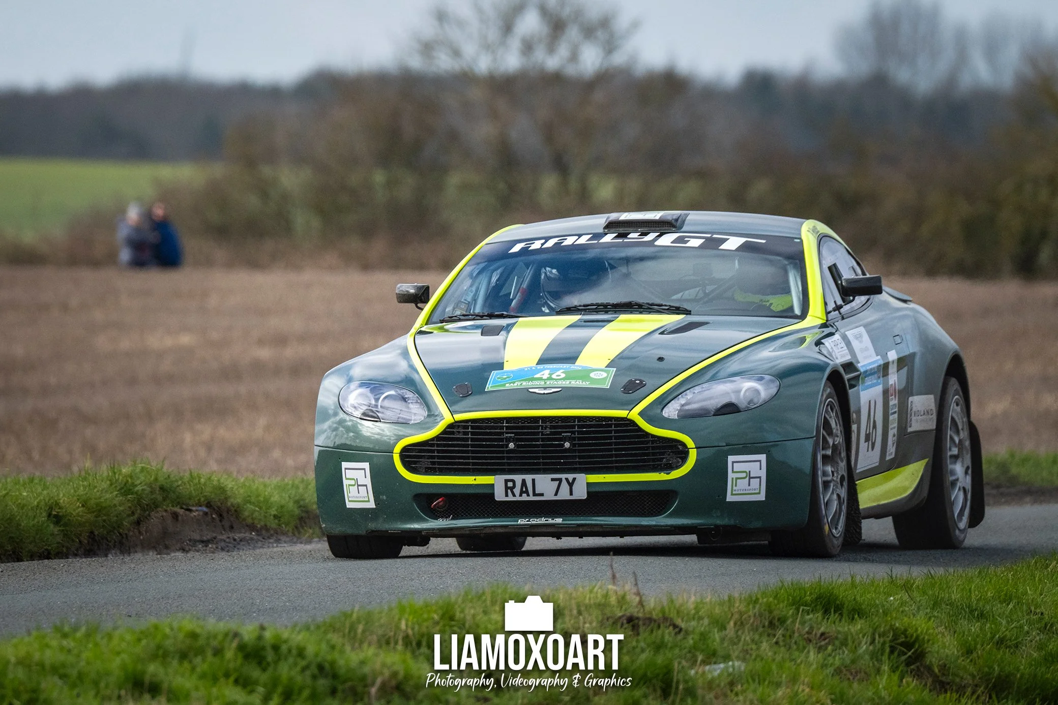 A green and black rally car on a dirt road, with a blurred background of trees and two people watching in the distance.