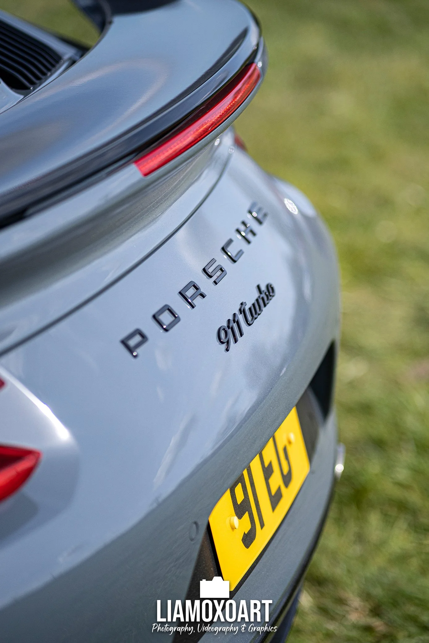 Close-up of the rear of a silver Porsche 911 Turbo with a yellow license plate, showing part of the car's model badge and sleek tail light, on a grassy area.