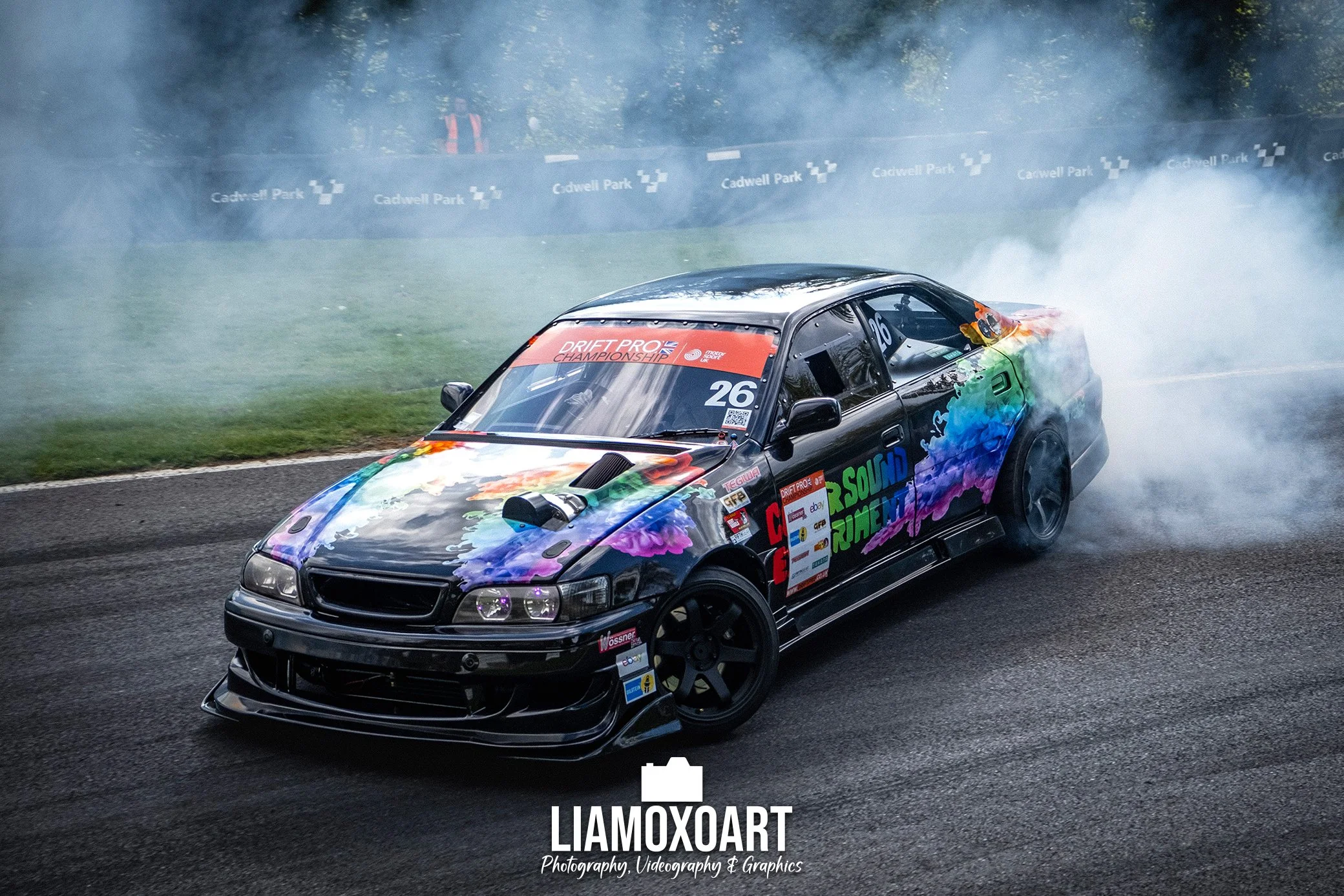 A black sports car with colorful paint splashes is drifting on a race track, creating smoke around its tires during a drifting competition at Cadwell Park. The car has sponsor stickers and a number 26 on the windshield.