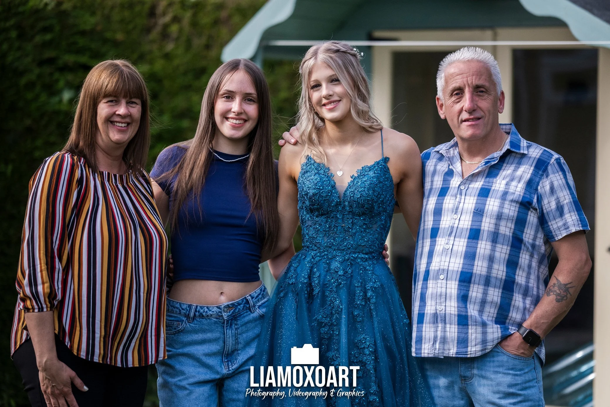 A family of four standing outdoors in front of a green hedge and a house, smiling at the camera. The family consists of a woman with brown hair wearing a striped shirt, a young woman in a blue top, a young woman in a blue dress with blonde hair, and 