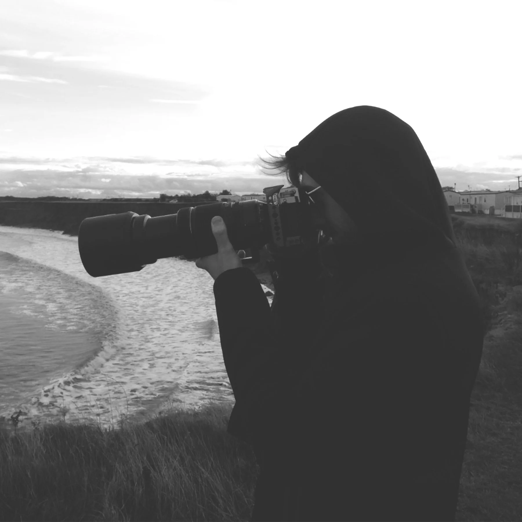 A person in a hoodie and sunglasses taking a photograph with a large camera lens near a body of water on a cloudy day.