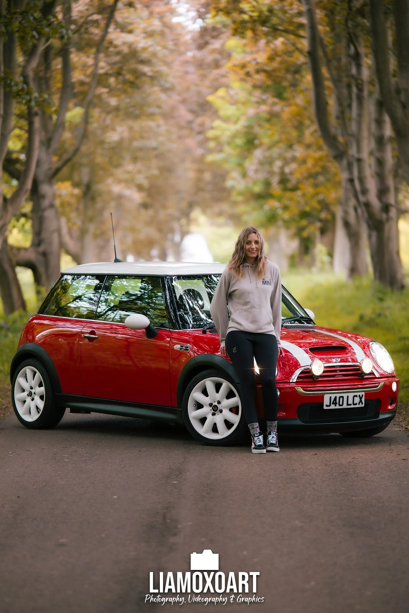 A smiling young woman with wavy blonde hair, wearing a light gray hoodie and black pants, standing in front of a red Mini Cooper car parked on a paved road surrounded by tall trees with autumn leaves.