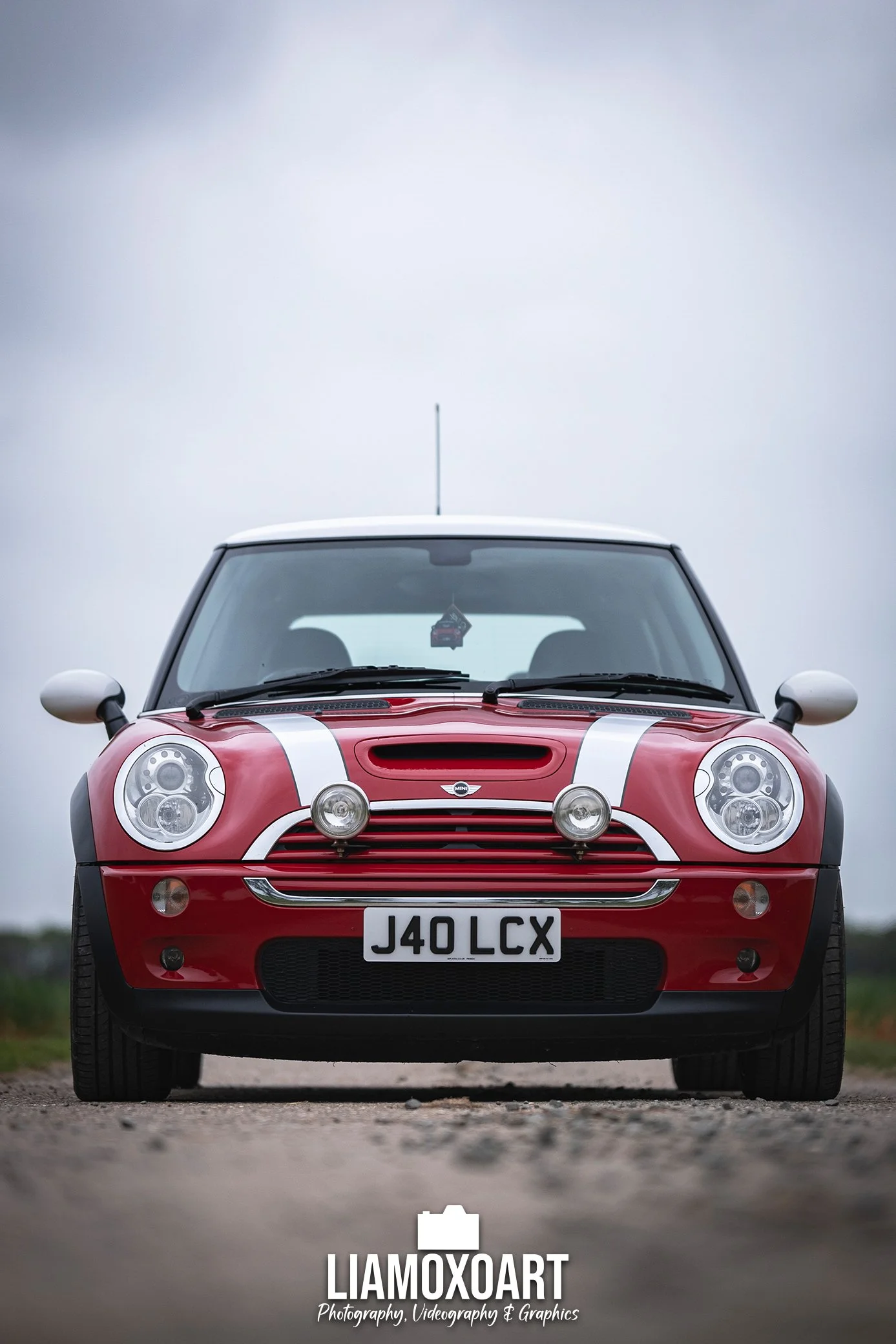 Front view of a red Mini Cooper with racing stripes, license plate J40 LCX, parked on a gravel road against a cloudy sky background.