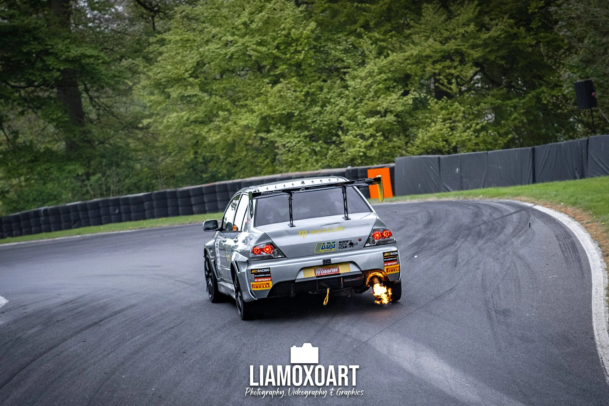 A silver race car on a winding track with a large rear wing, surrounded by green trees, emitting flames from the exhaust in a racing environment.