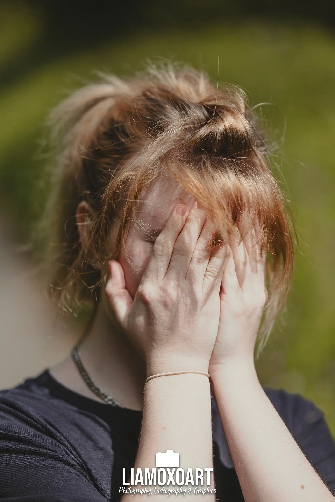 A person with red hair covering their face with their hands, wearing a black top, jewelry, and a bracelet, outdoors with blurred green background.