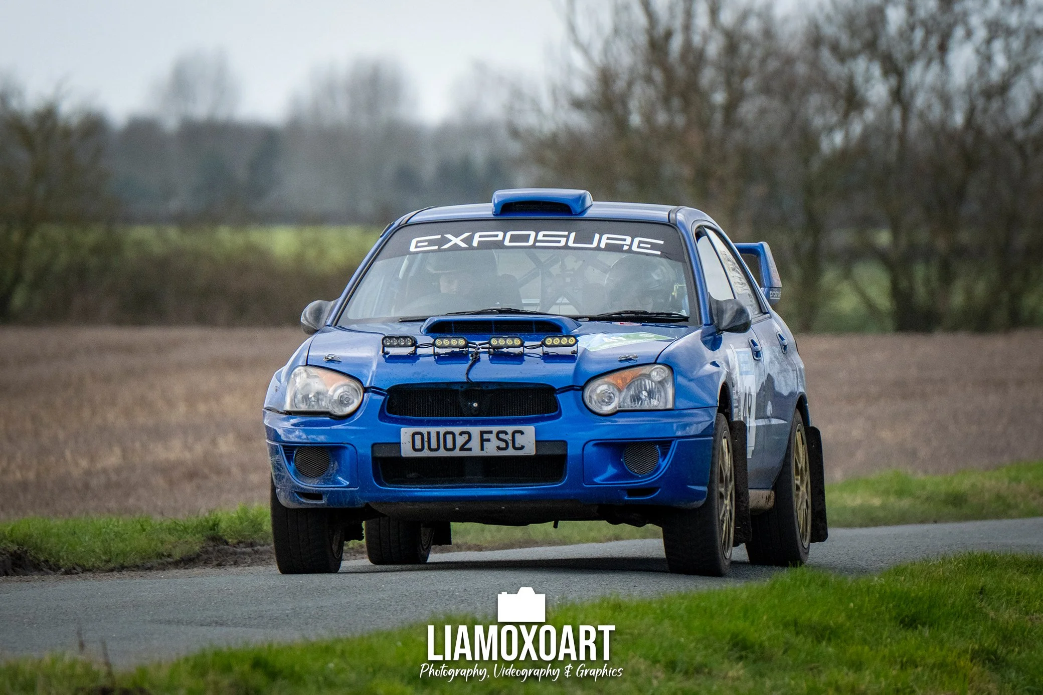 A blue rally car driving on a rural road with trees in the background.