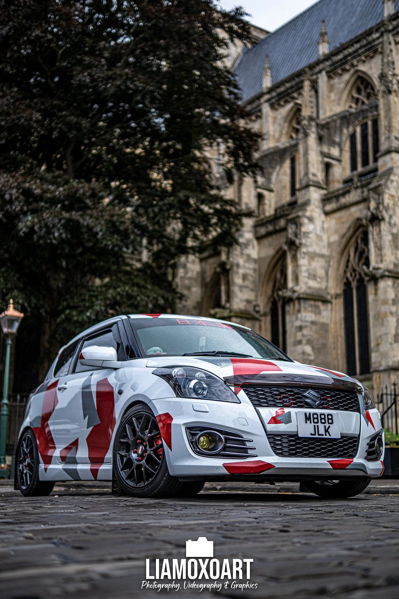 A white sports car with red and black camouflage graphics parked on cobblestone street in front of a historic stone church with gothic architecture and a dark tree.
