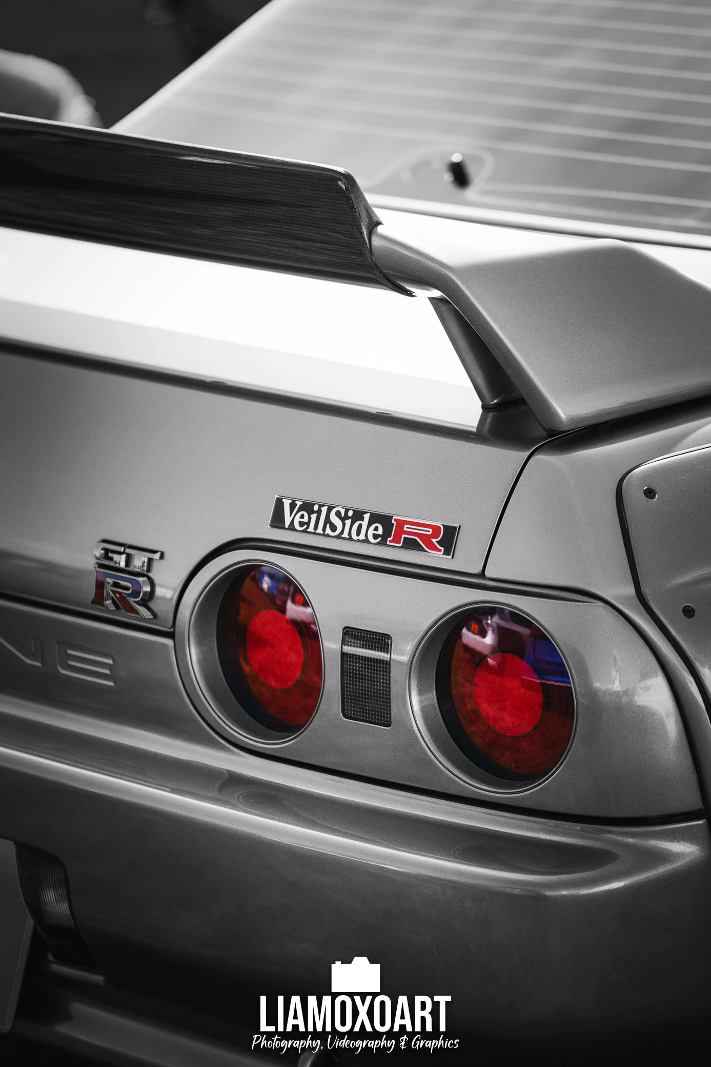 Close-up of the rear of a silver Nissan GT-R car, featuring a black rear wing spoiler and round taillights. The image showcases the car's branding and a branded watermark. Black-and-white photography emphasizes the car's design details.