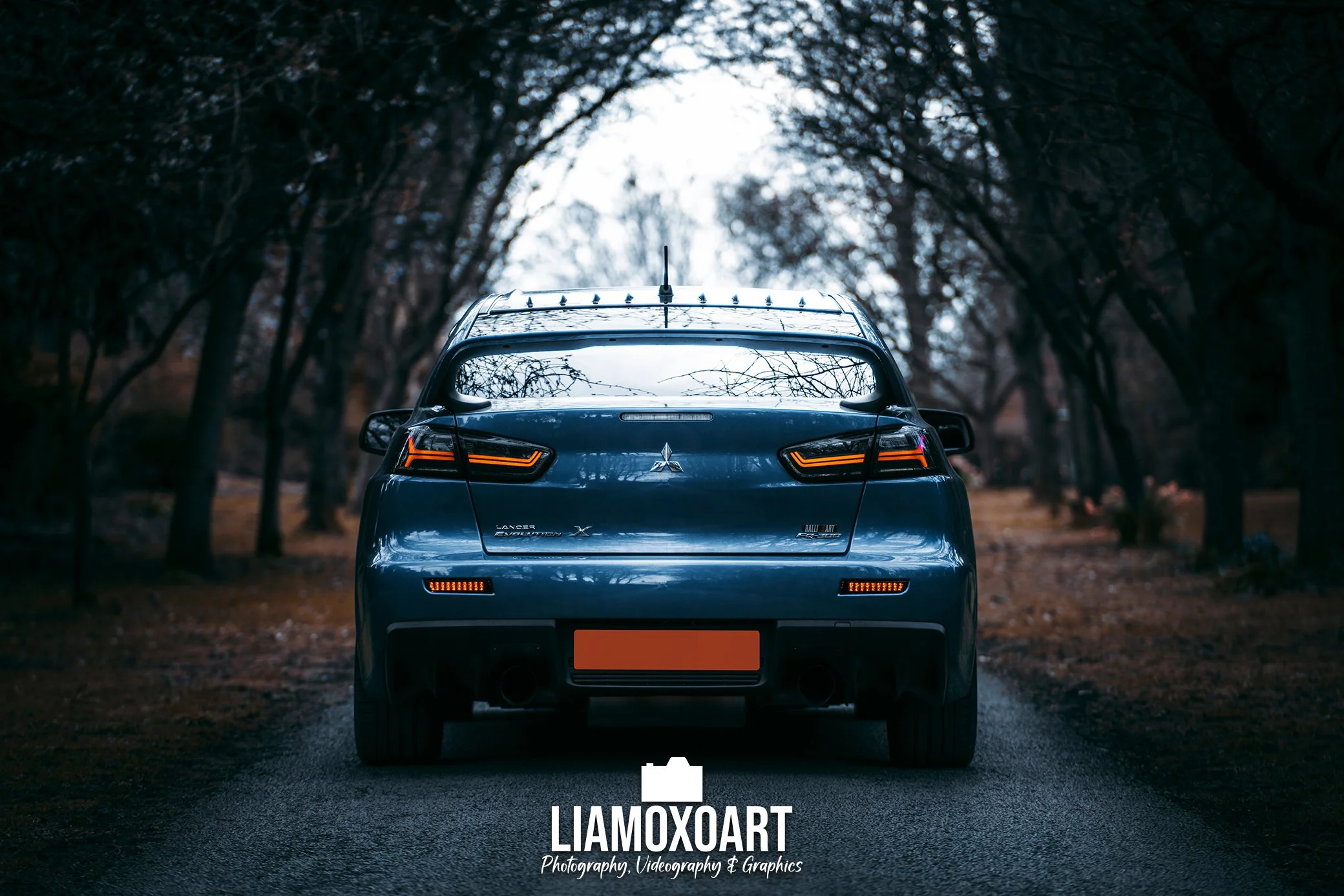 Rear view of a dark blue Mitsubishi Lancer Evolution X parked on a road surrounded by trees with fall foliage, taken during dusk.