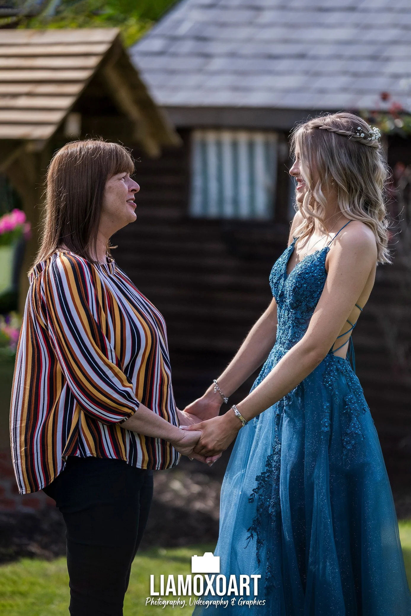 A woman with a floral tiara in a blue dress holding hands with another woman in a striped blouse and dark pants, in a garden with a wooden house in the background.