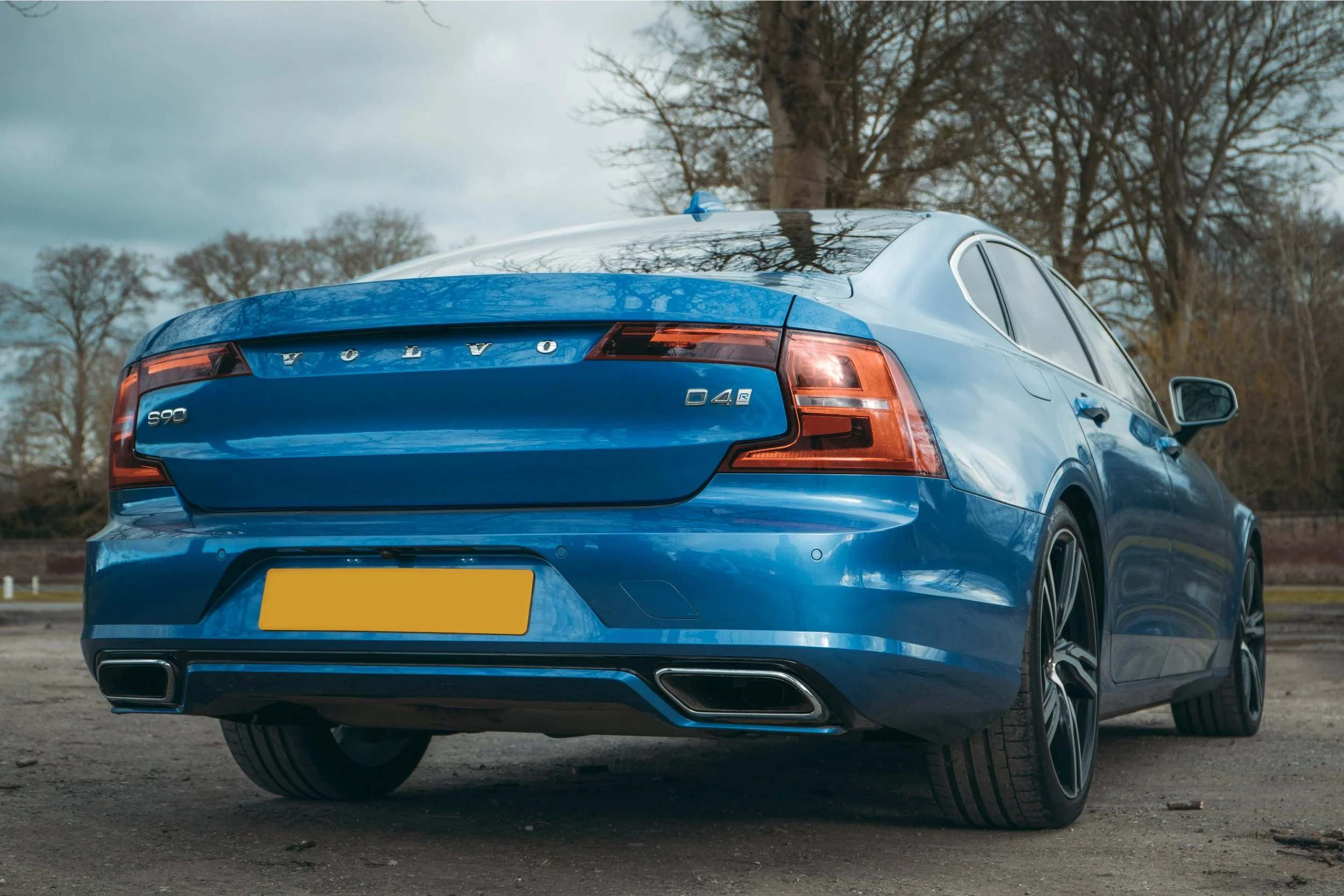 Blue Volvo S90 sedan parked outdoors on a cloudy day with trees in the background.