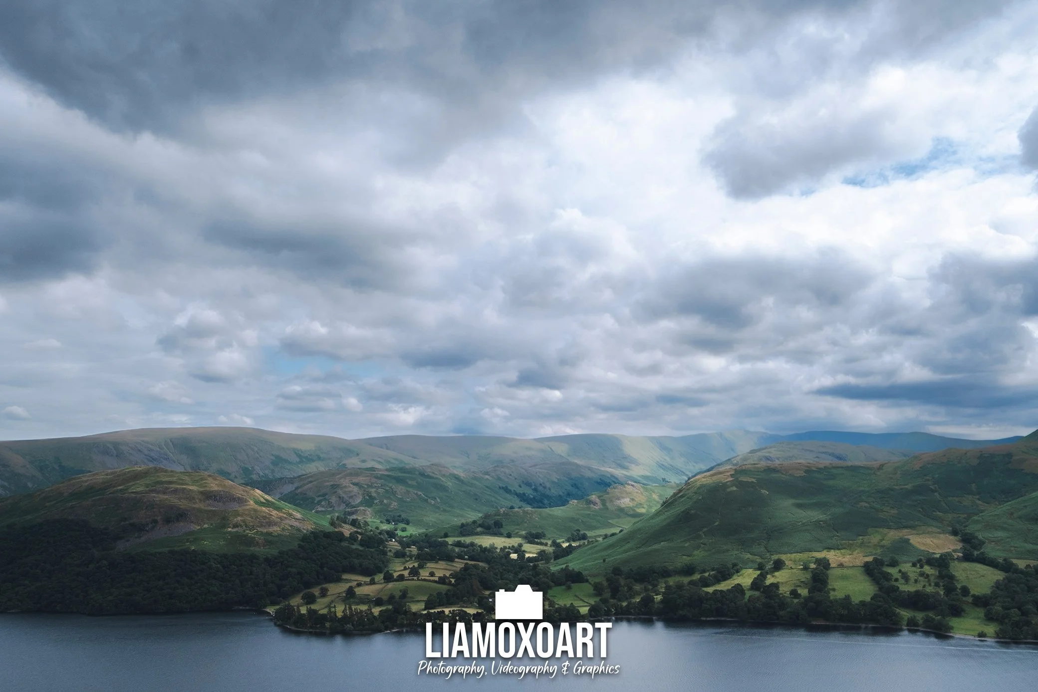 Scenic view of rolling green hills and mountains with a lake in the foreground under a cloudy sky.