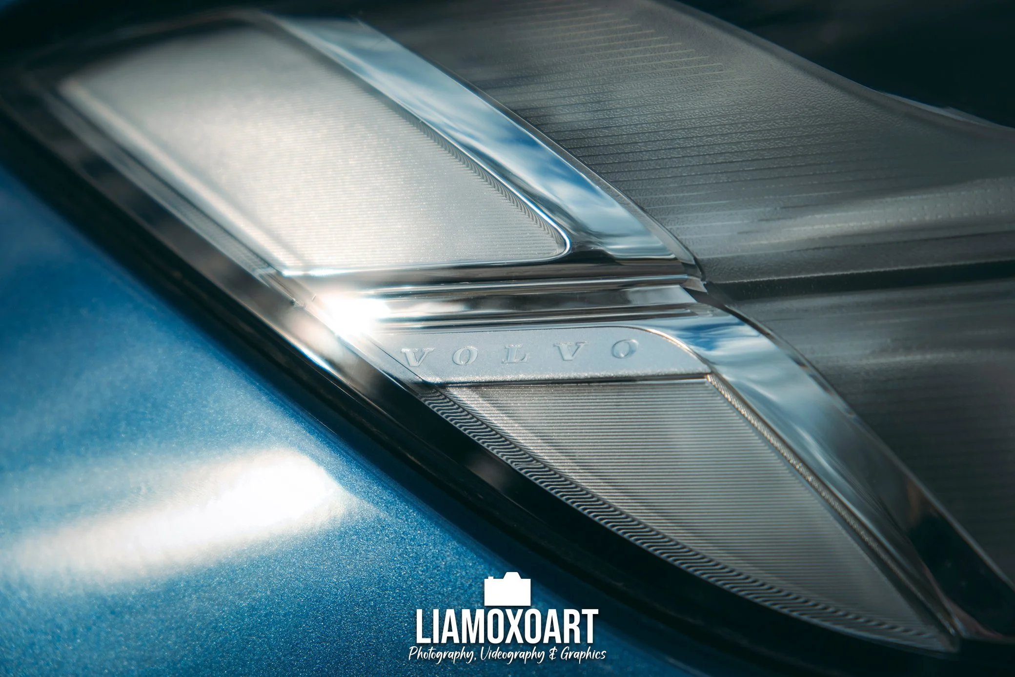 Close-up of a car headlight with the brand name VOLVO engraved on it, set against a blue painted vehicle body.