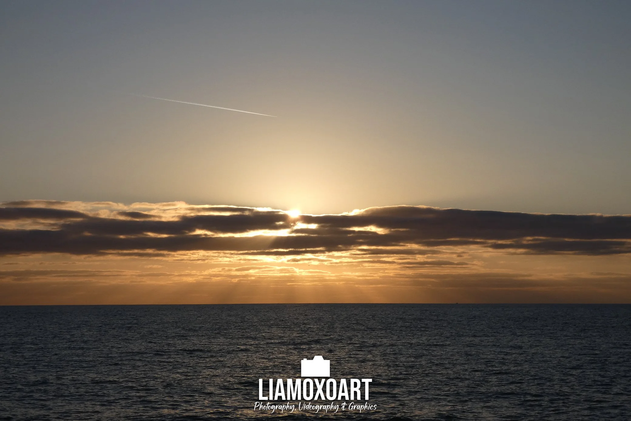 Sunset over the ocean with a mostly clear sky and some clouds near the horizon. The sun is partially obscured by clouds, casting a warm glow across the water.