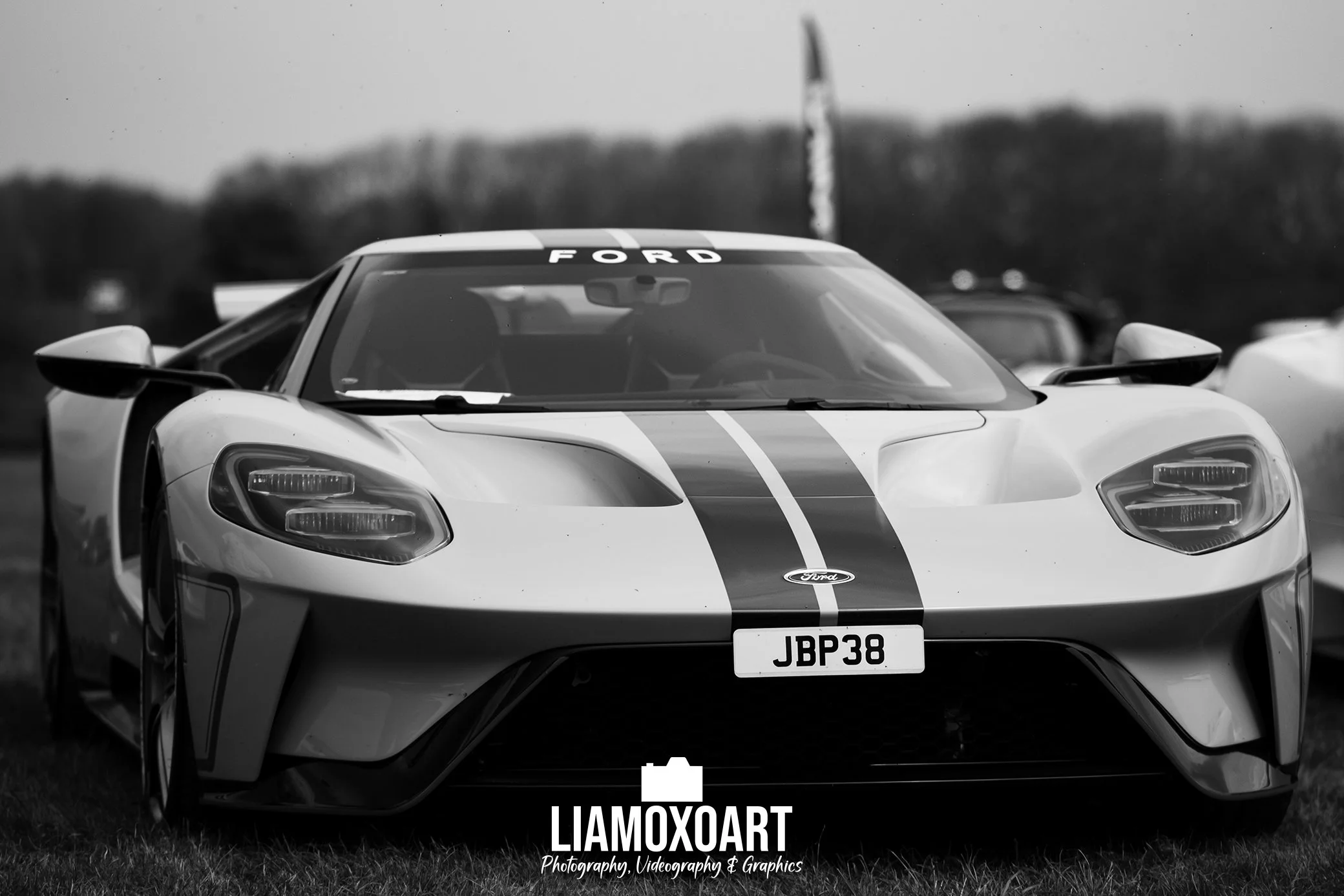 A black and white photograph of a Ford sports car, possibly a GT model, with a prominent front bumper, sharp headlight design, and a racing stripe running from the hood to the windshield. The car's license plate reads JBP38, and the windshield has th
