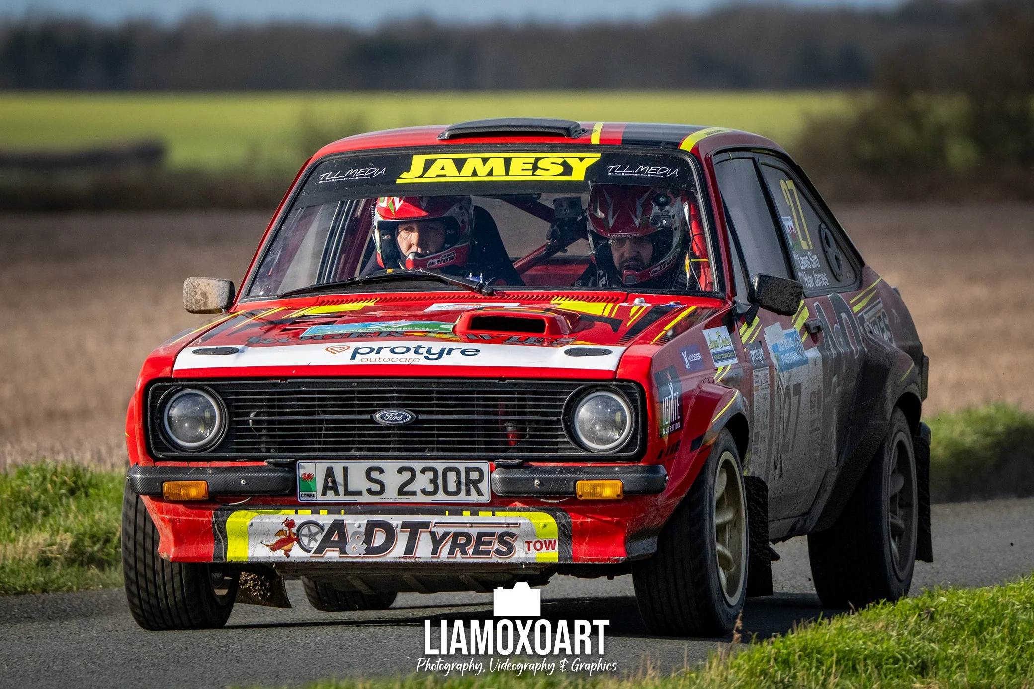 Red rally car with racing helmets inside, driving on a dirt path.