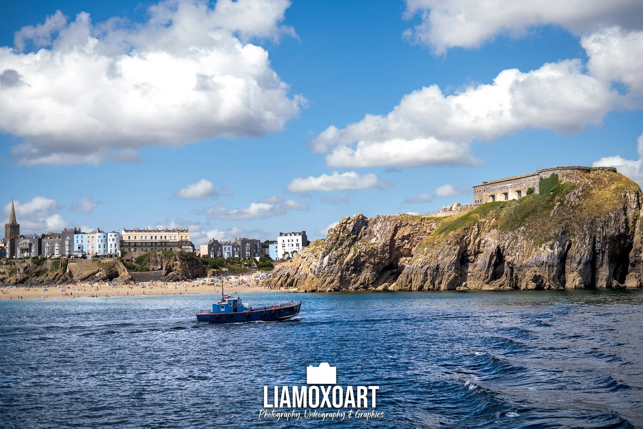 A coastal scene with a sandy beach filled with people, a boat on the water, a rocky cliff with a historical building on top, and a blue sky with fluffy white clouds.
