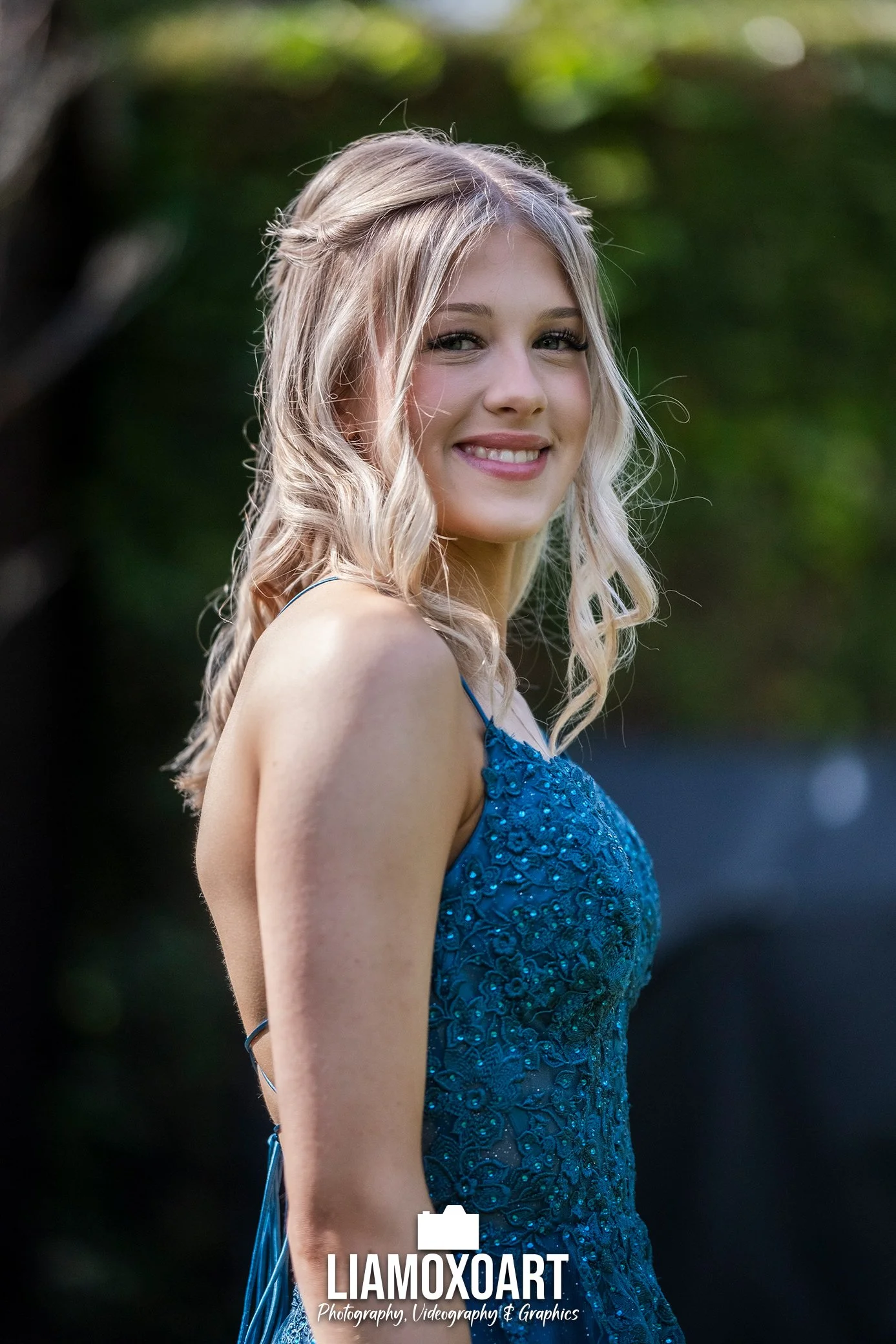 A young woman with blonde hair styled in loose waves, smiling and looking at the camera, wearing a blue, sleeveless, embroidered dress outdoors with a blurred green background.