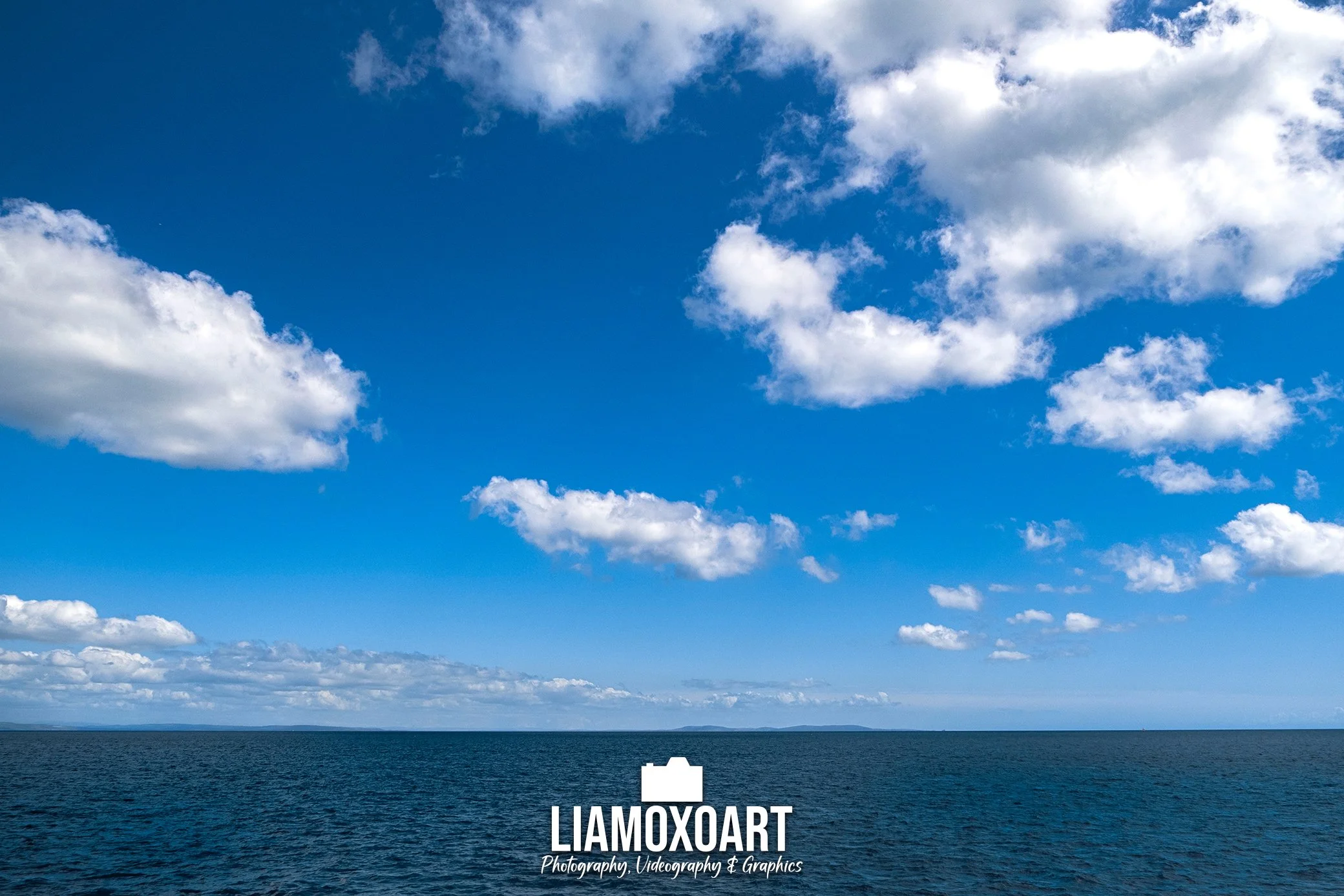 A vast body of water, possibly an ocean or large lake, with a distant horizon. Above, a bright blue sky filled with scattered white clouds.