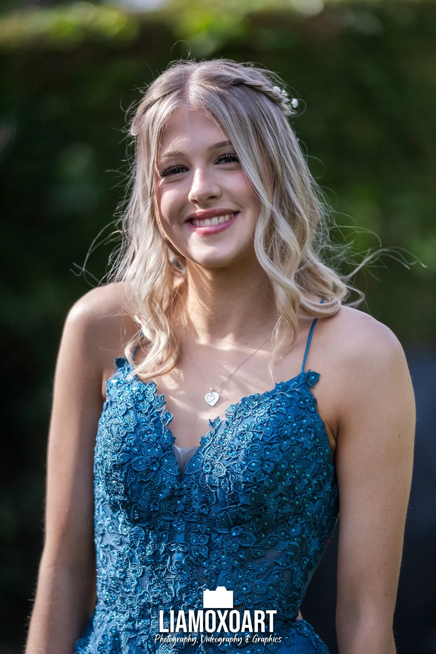 A young woman with blonde, wavy hair dressed in a blue, embroidered dress, smiling outdoors with a blurred green background.