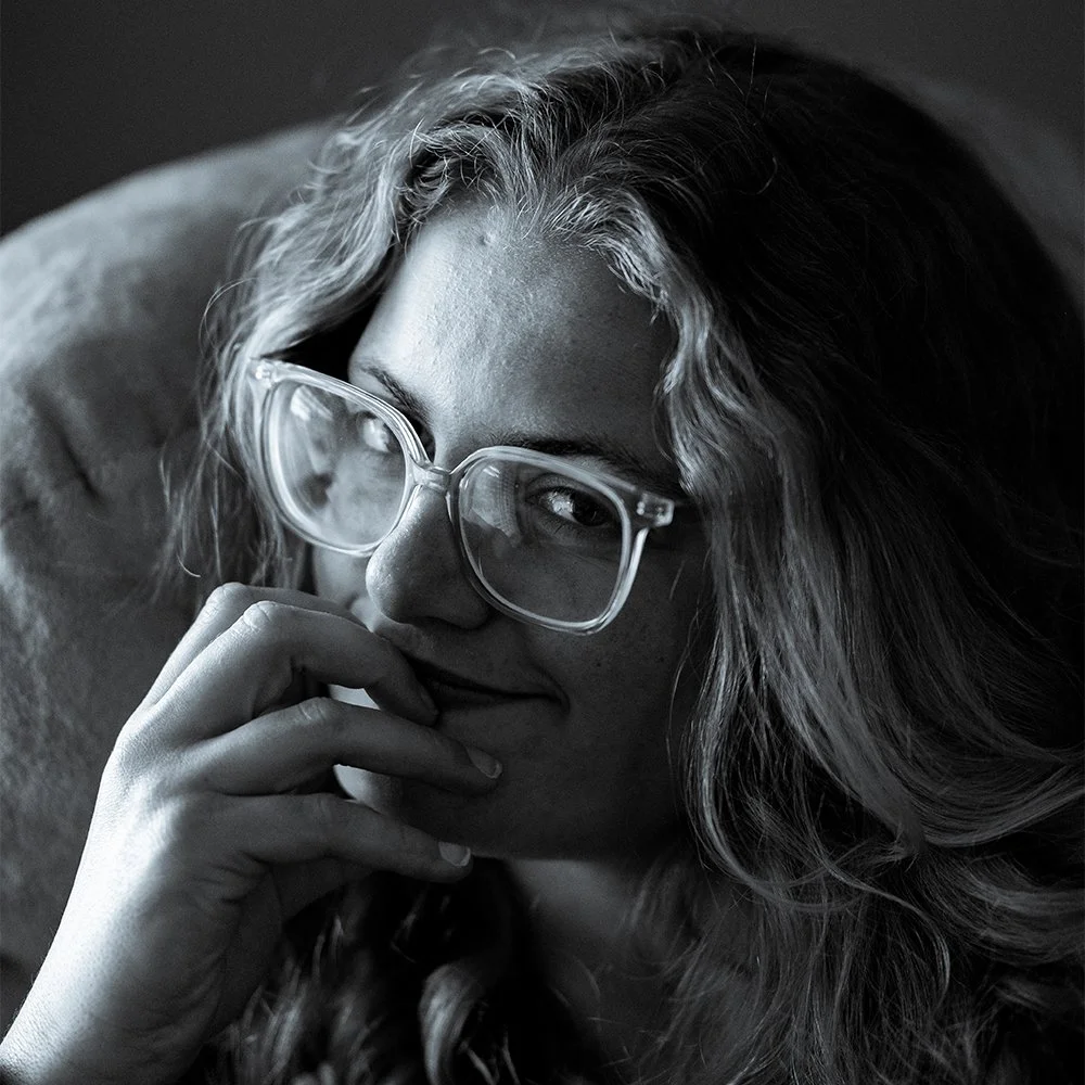 Black and white portrait of a woman with wavy hair wearing glasses, resting her chin on her hand, looking at the camera.