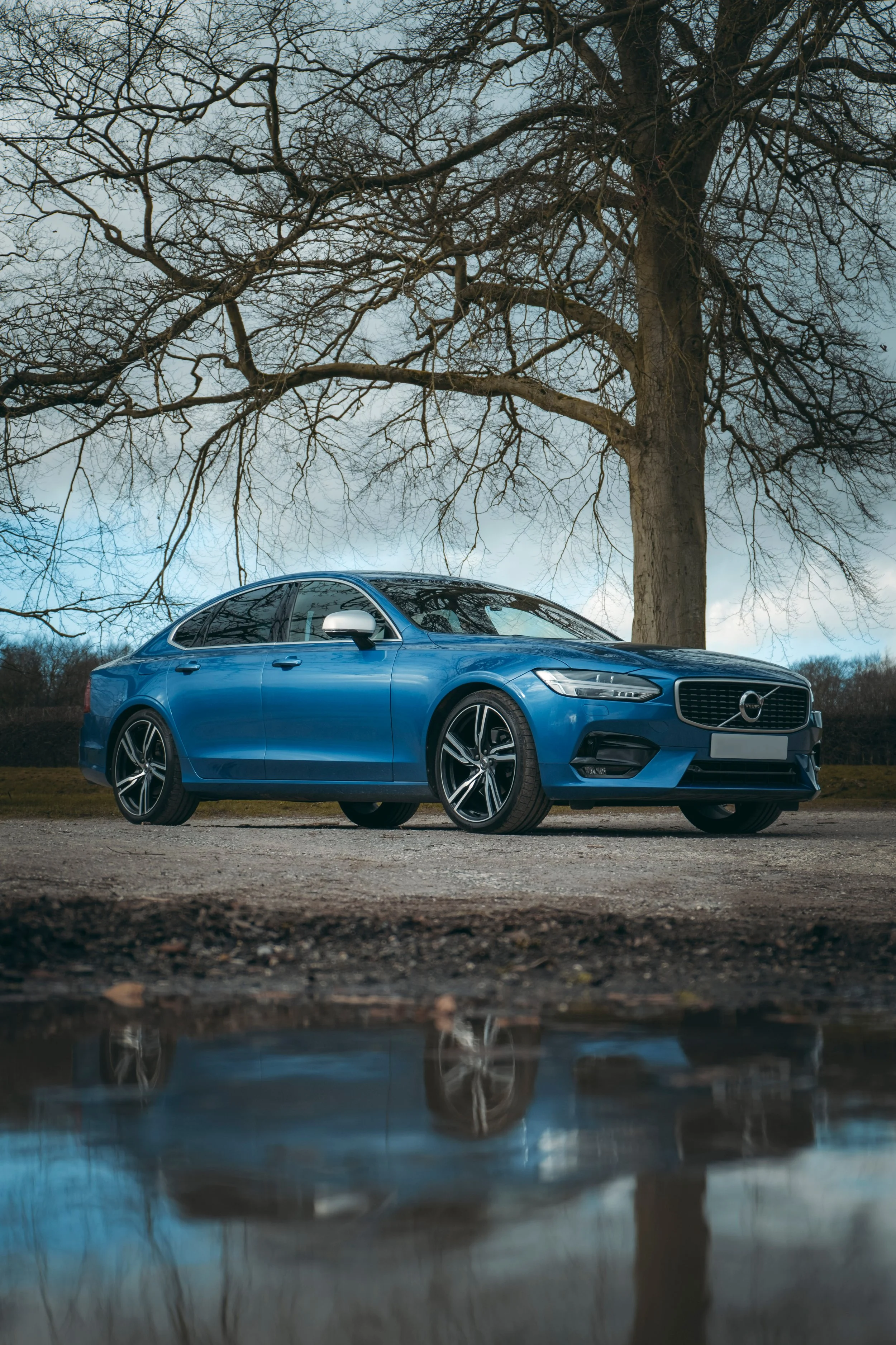 Blue sedan car parked on a gravel surface under leafless tree, with its reflection visible in a small puddle on the ground.
