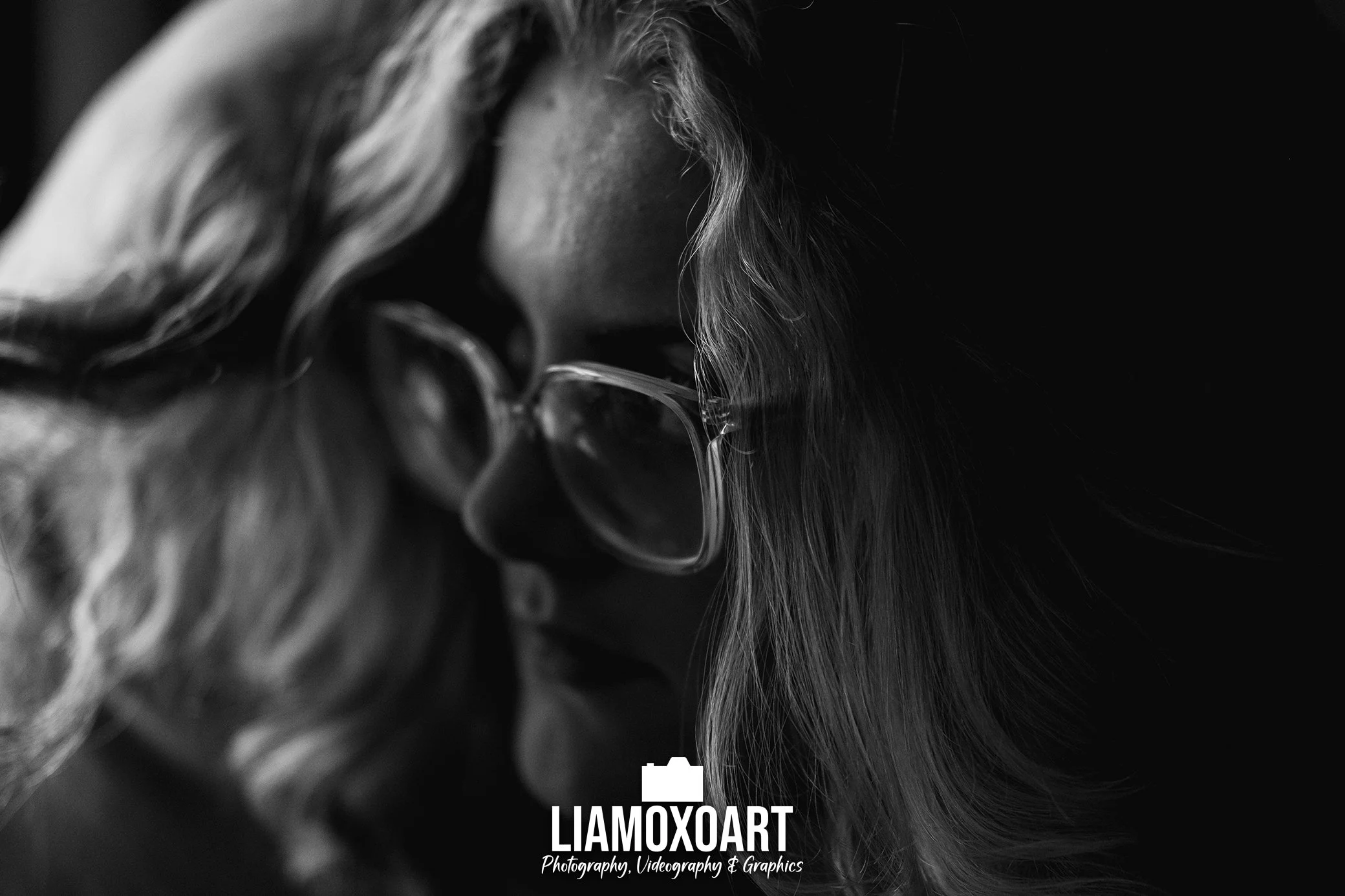 Close-up black and white photograph of a person with long, curly hair and glasses, focused on their face.