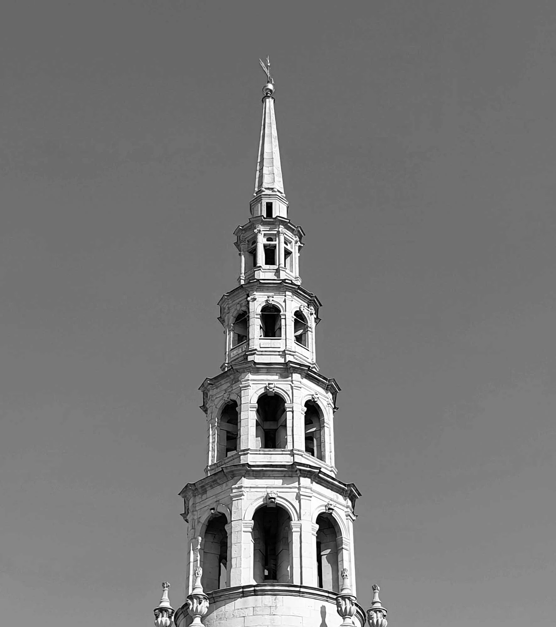 Black and white photo of a tall, ornate church steeple with multiple tiers and arched openings, topped with a weather vane and a figure at the very top.