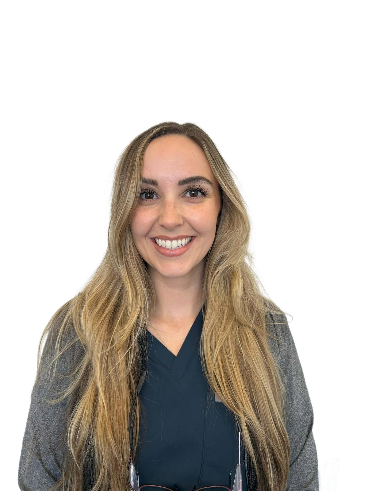 Young woman with long wavy hair smiling, wearing navy scrubs and sneakers, standing in front of a glass door with a black logo, in a modern office or clinic setting.