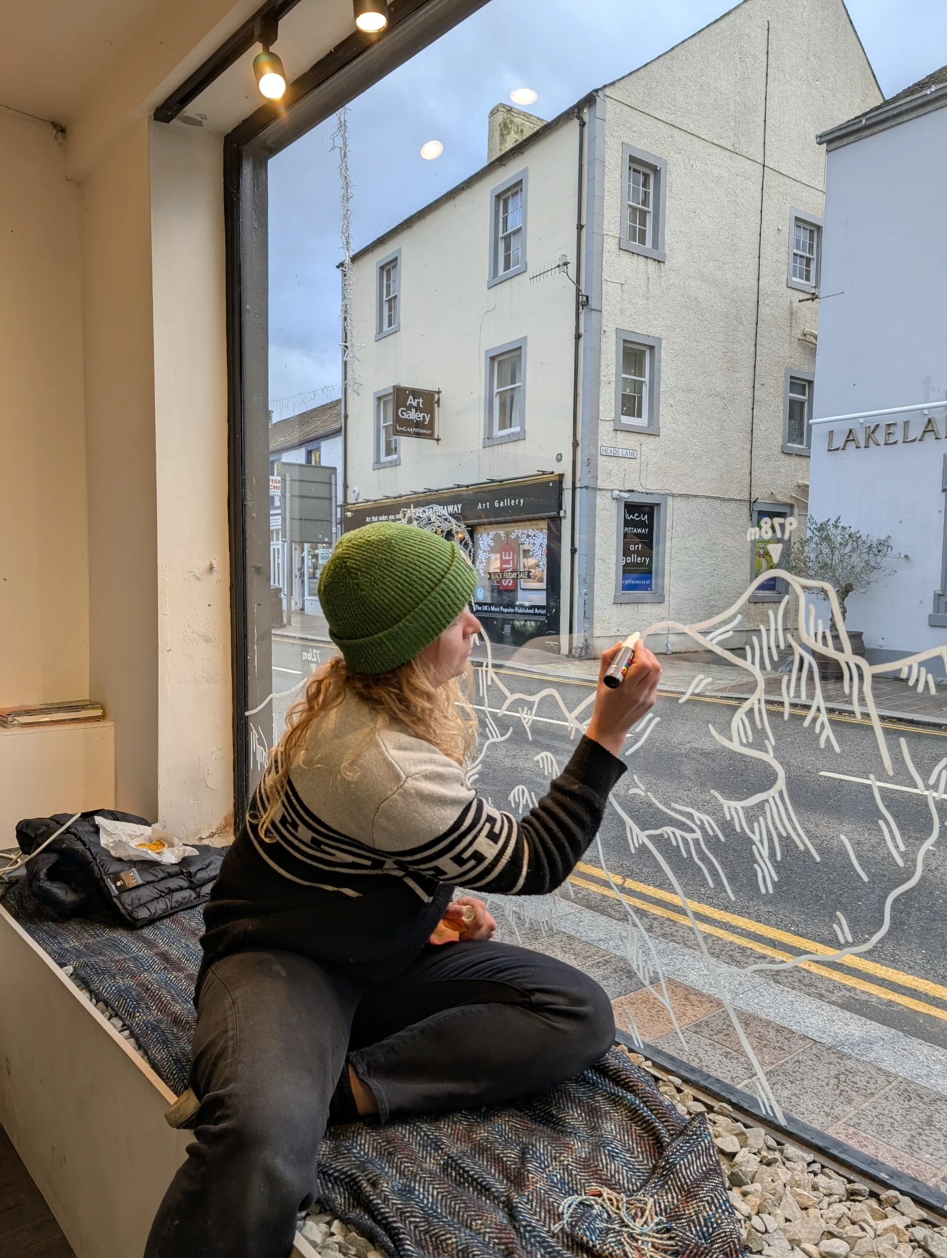 A woman in a green beanie and black and white sweater sitting on a window seat, drawing on the glass with a white marker. Outside the window are buildings and signs, including an art gallery.