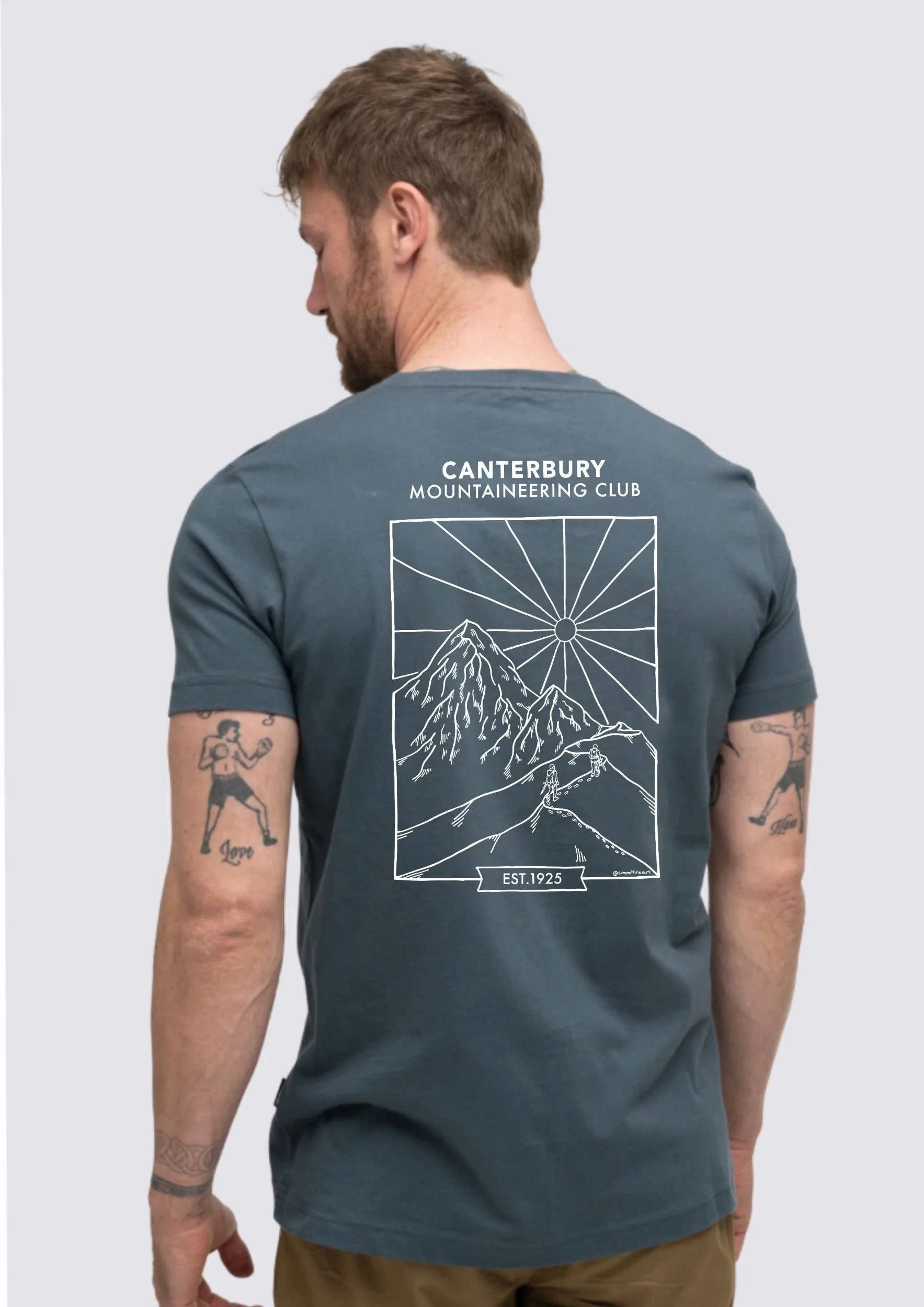 Back view of a man wearing a blue T-shirt with a mountain landscape and sun design, with text indicating 'Canterbury Mountaineering Club Est.1925', against a plain light background.