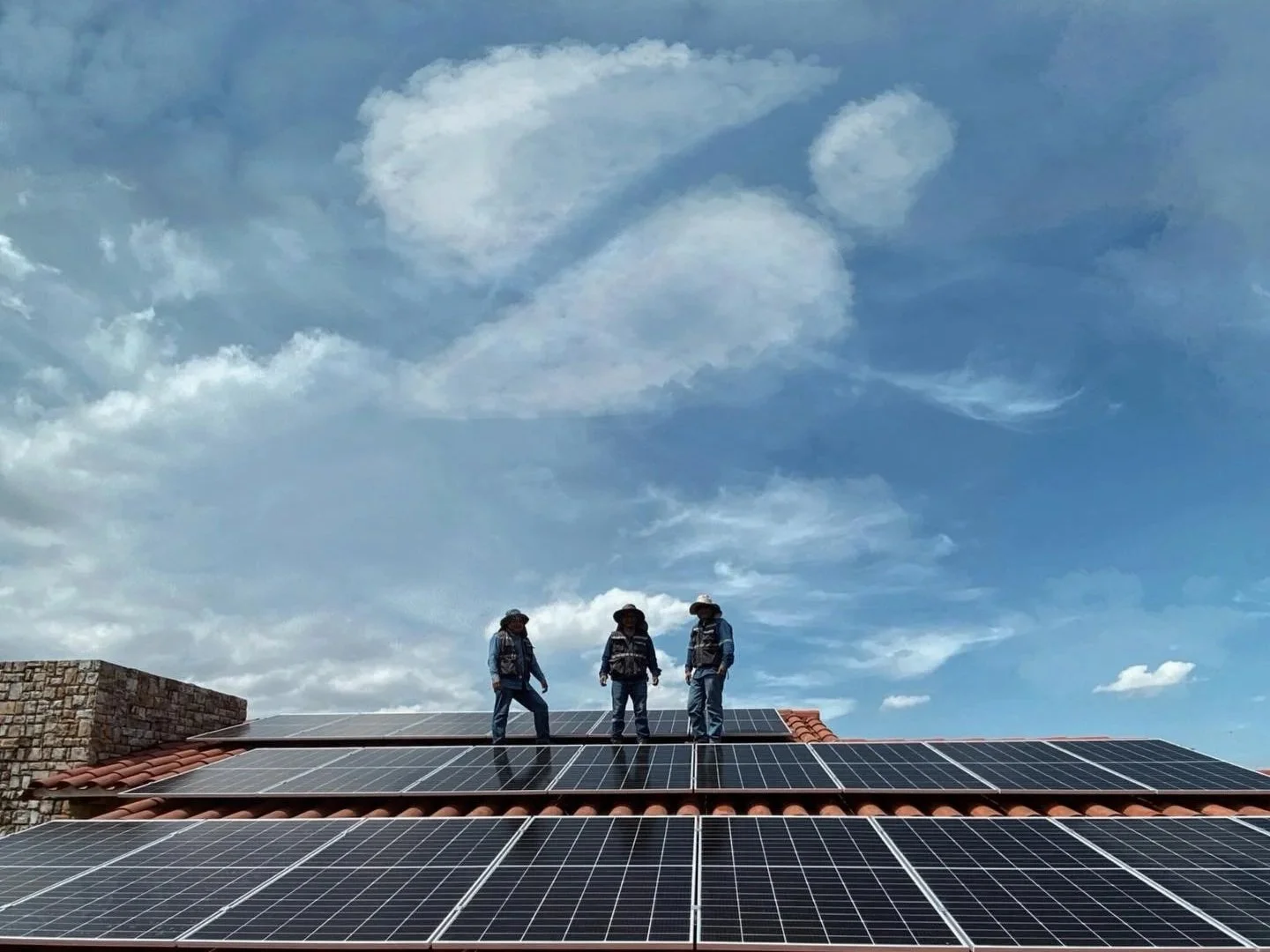 Tres trabajadores en el techo instalando paneles solares contra un cielo azul con nubes.