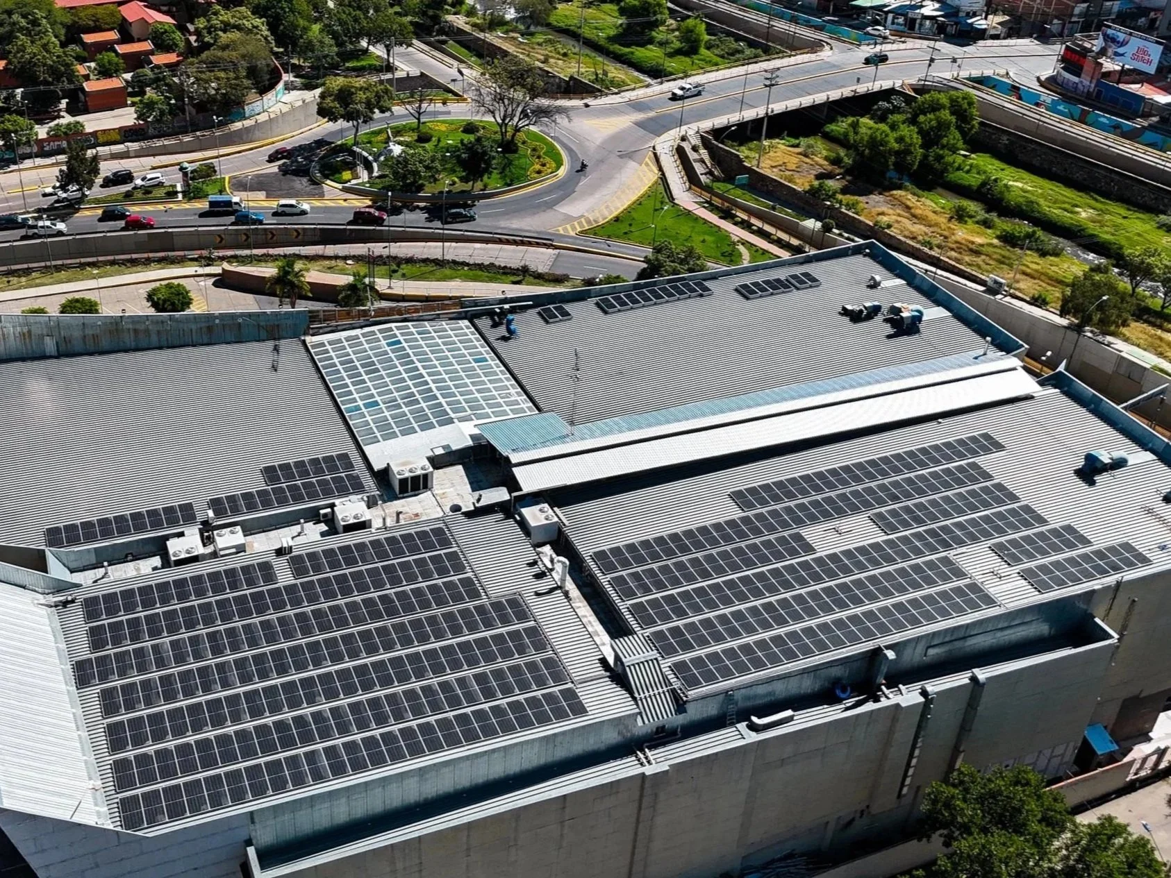 Vista aérea de techos de un edificio con paneles solares instalados, en un entorno urbano con calles, vehículos y áreas verdes cercanas.