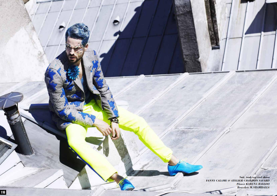 Un homme assis sur un toit en métal, portant un costume avec des chaussures bleues et des pantalons jaunes fluorescent, avec une veste à motifs et des accessoires de mode.