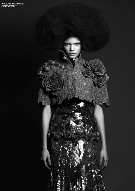 Une femme porte une robe et un manteau ornés de rosettes, avec une coiffure volumineuse et bouffante, en noir et blanc.