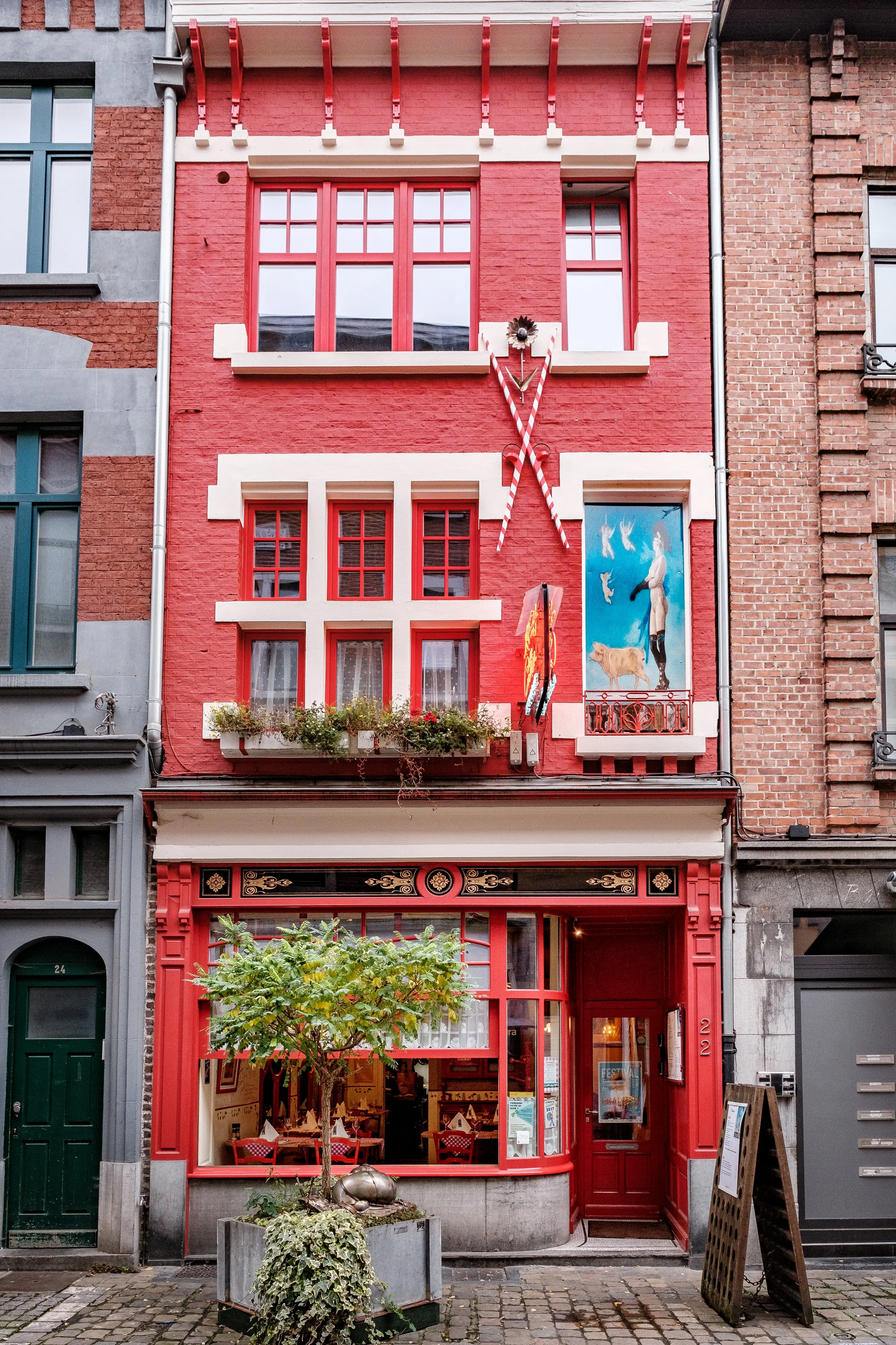 Facade d'un restaurant coloré rouge avec grande fenêtre au rez-de-chaussée, petite fenêtre à gauche, et fenêtres plus grandes à l'étage, décorée de plantes, avec un tableau peint à droite, dans une rue pavée.