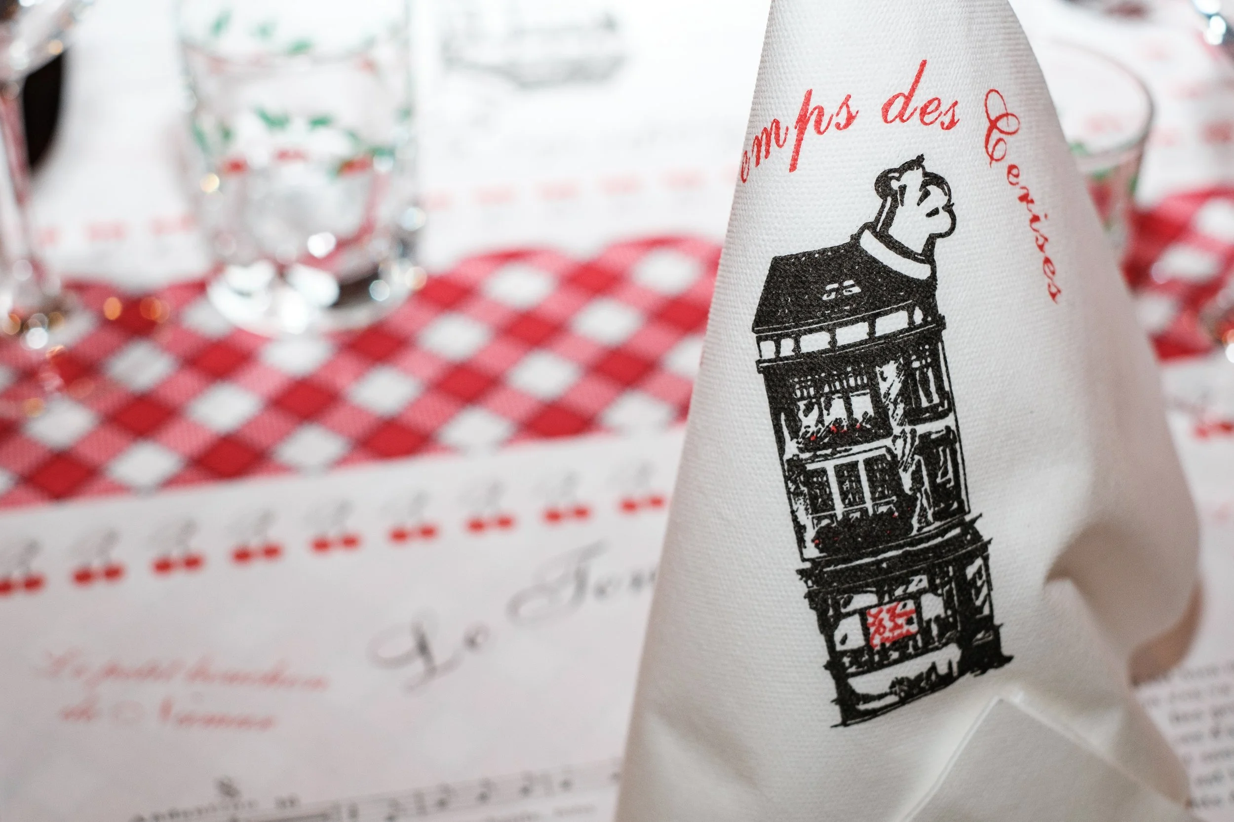 Une serviette en tissu blanc avec une illustration en noir d'une horloge à quatre étages et des inscriptions en rouge, posée à côté d'une table à nappes rouge et blanche à carreaux.