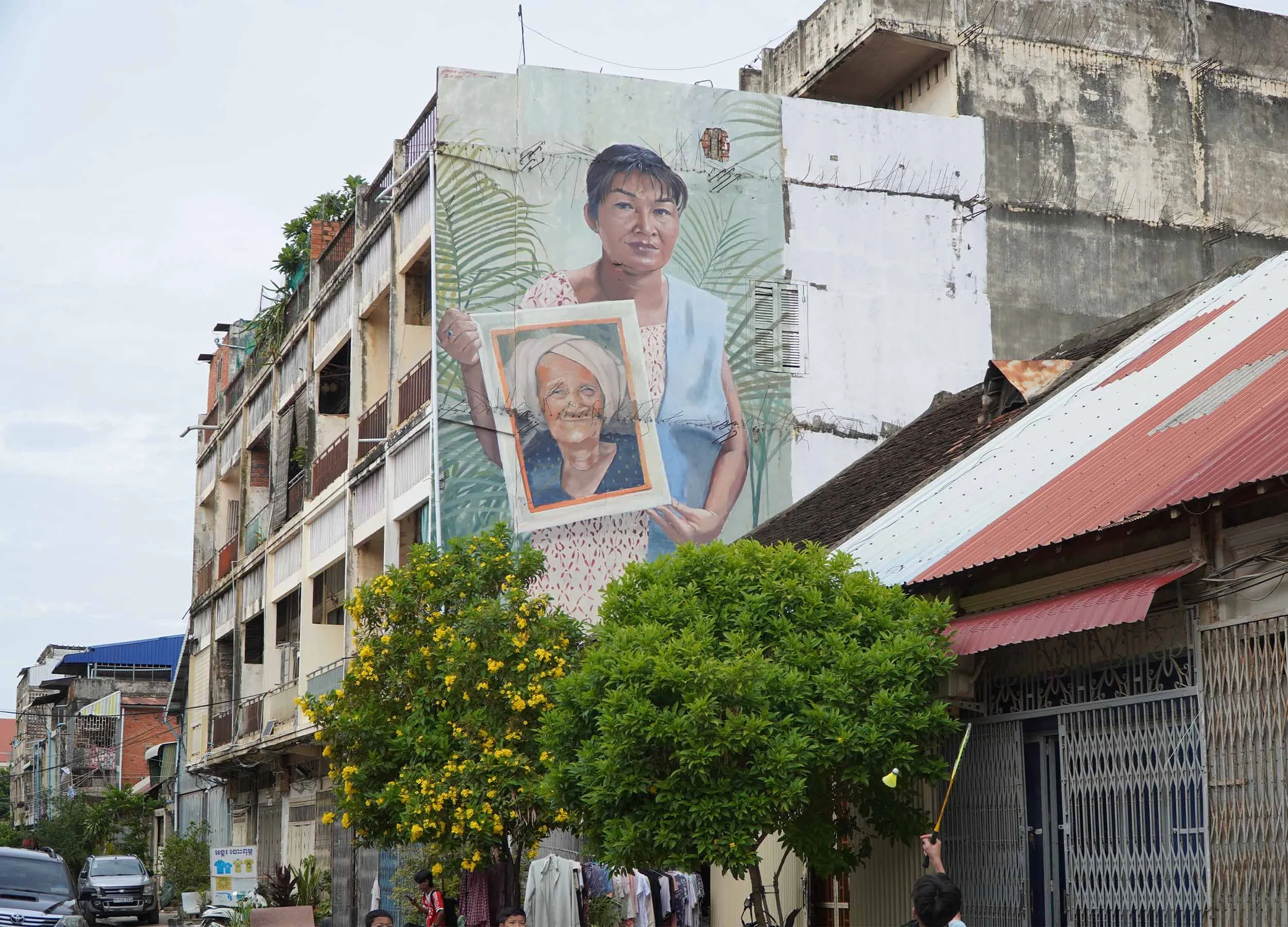 Large mural of a woman holding a framed picture of her grandma on a building in an urban area. This is on street 1.5, Battambang city