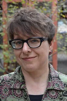 A person with short gray hair and glasses outdoors, wearing a patterned jacket.