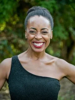 Portrait of a smiling woman outdoors with green trees in the background, wearing a black sleeveless top.