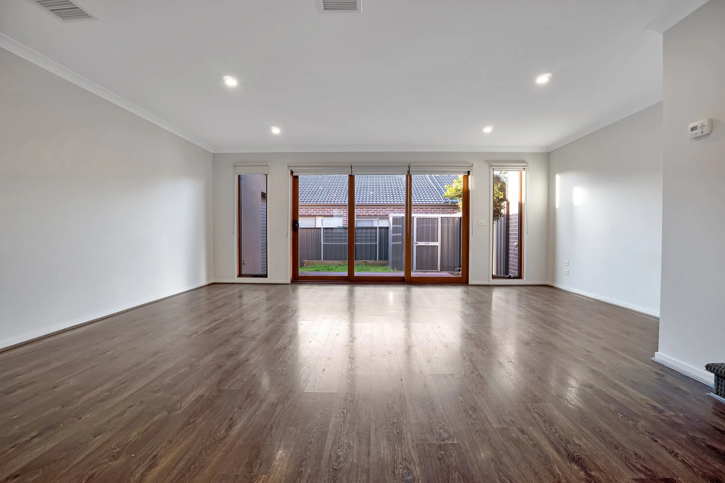 Empty living room with hardwood floors, white walls, and large glass sliding door leading to backyard. Two tall narrow windows on either side of sliding door. Bright natural light inside.