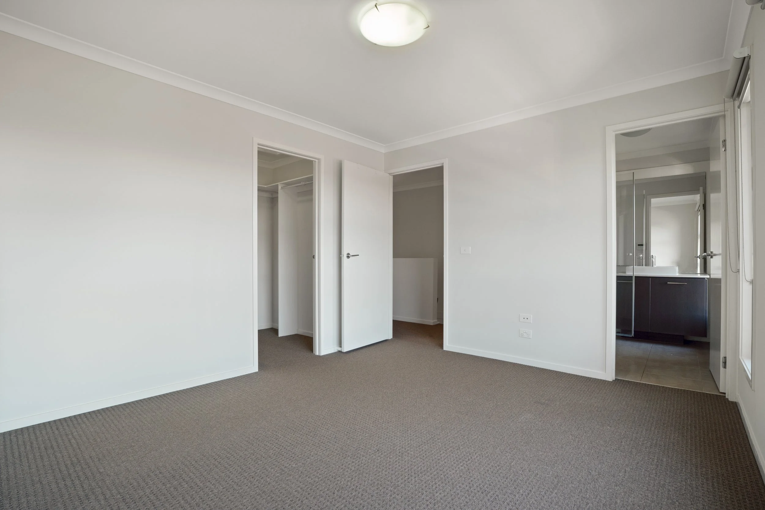 Empty bedroom with beige walls, brown carpet, two open doorways, and a view into a bathroom with a dark wood vanity and a white sink.