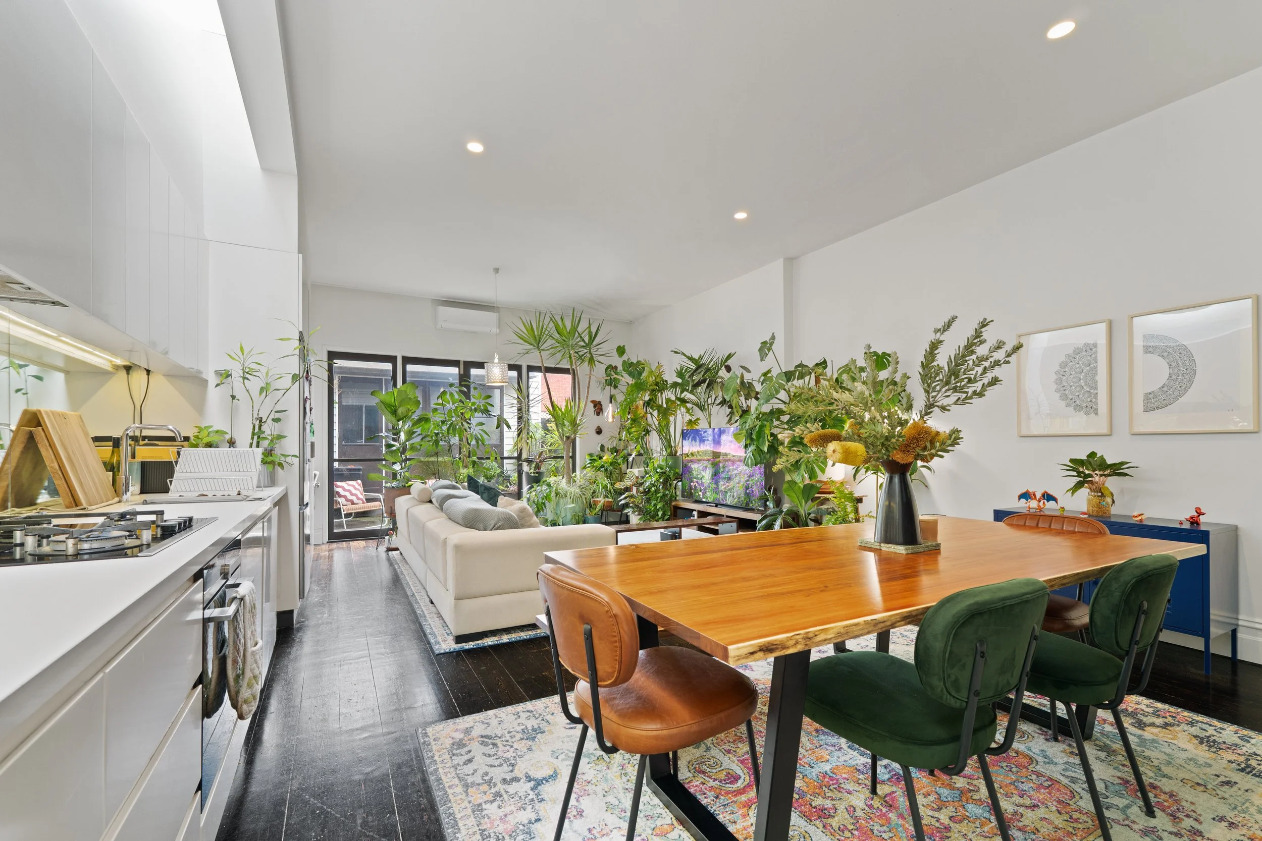 Open-concept living and dining area with a modern kitchen, a white sofa, a wooden dining table with four chairs, and numerous green indoor plants.