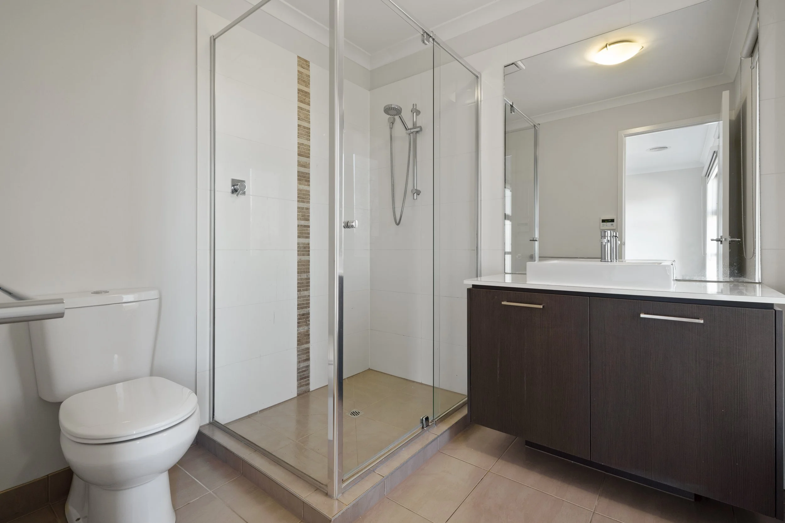Modern bathroom with a white toilet, a glass shower enclosure with tiled walls, a dark wood vanity with a sink, large mirror, and beige tiled flooring.