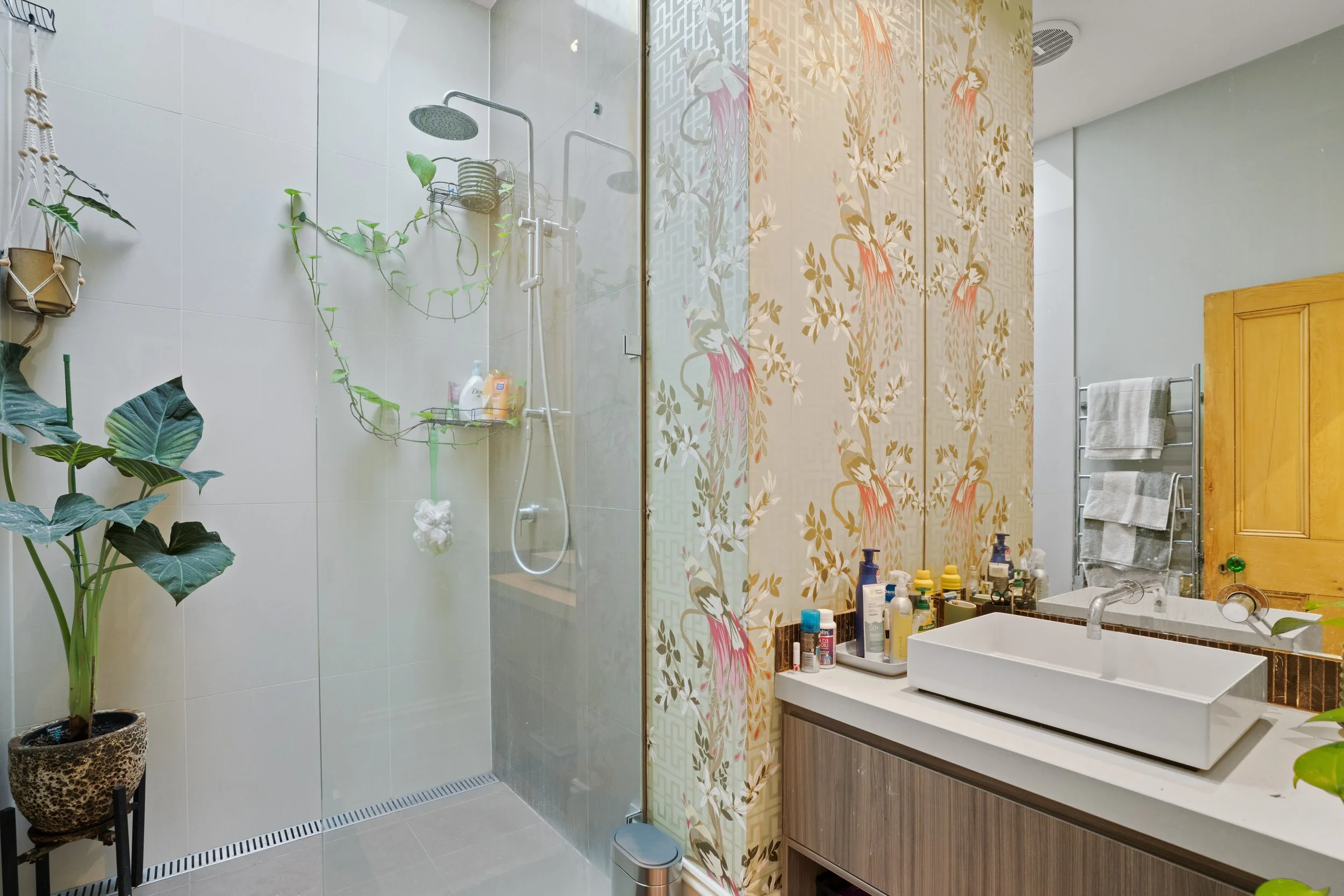 Modern bathroom with a walk-in shower and a vanity with a rectangular vessel sink. The shower has a glass partition, a rain showerhead, and some plants and toiletries inside. The vanity area has a mirror, an array of toiletries, and towels on a towel rack. There are decorative floral wallpaper and potted plants adding a touch of nature to the space.