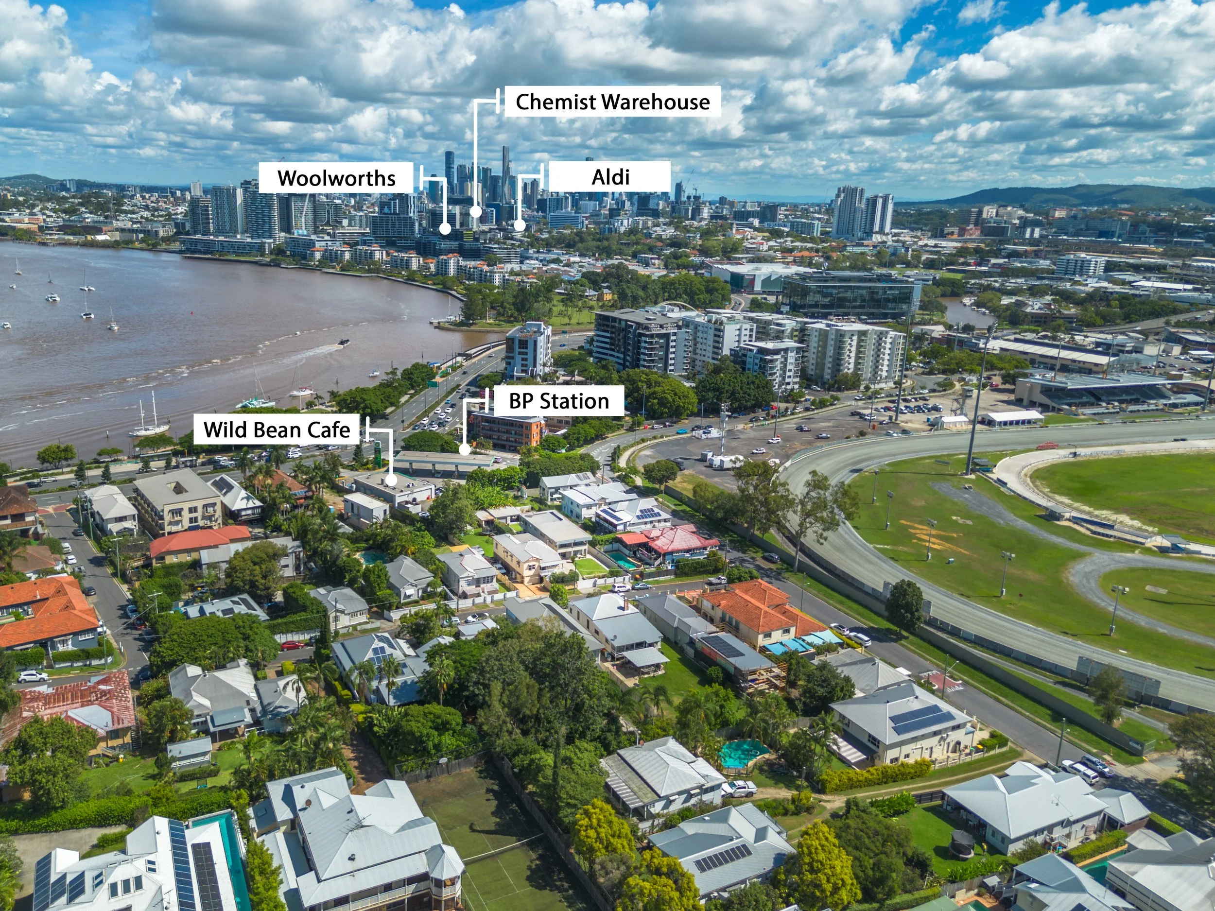 Aerial view of a city with labels indicating landmarks including Woolworths, Wild Bean Cafe, BP Station, Aldi, Chemist Warehouse, and a river with boats, with high-rise buildings in the background and a residential neighborhood in the foreground.