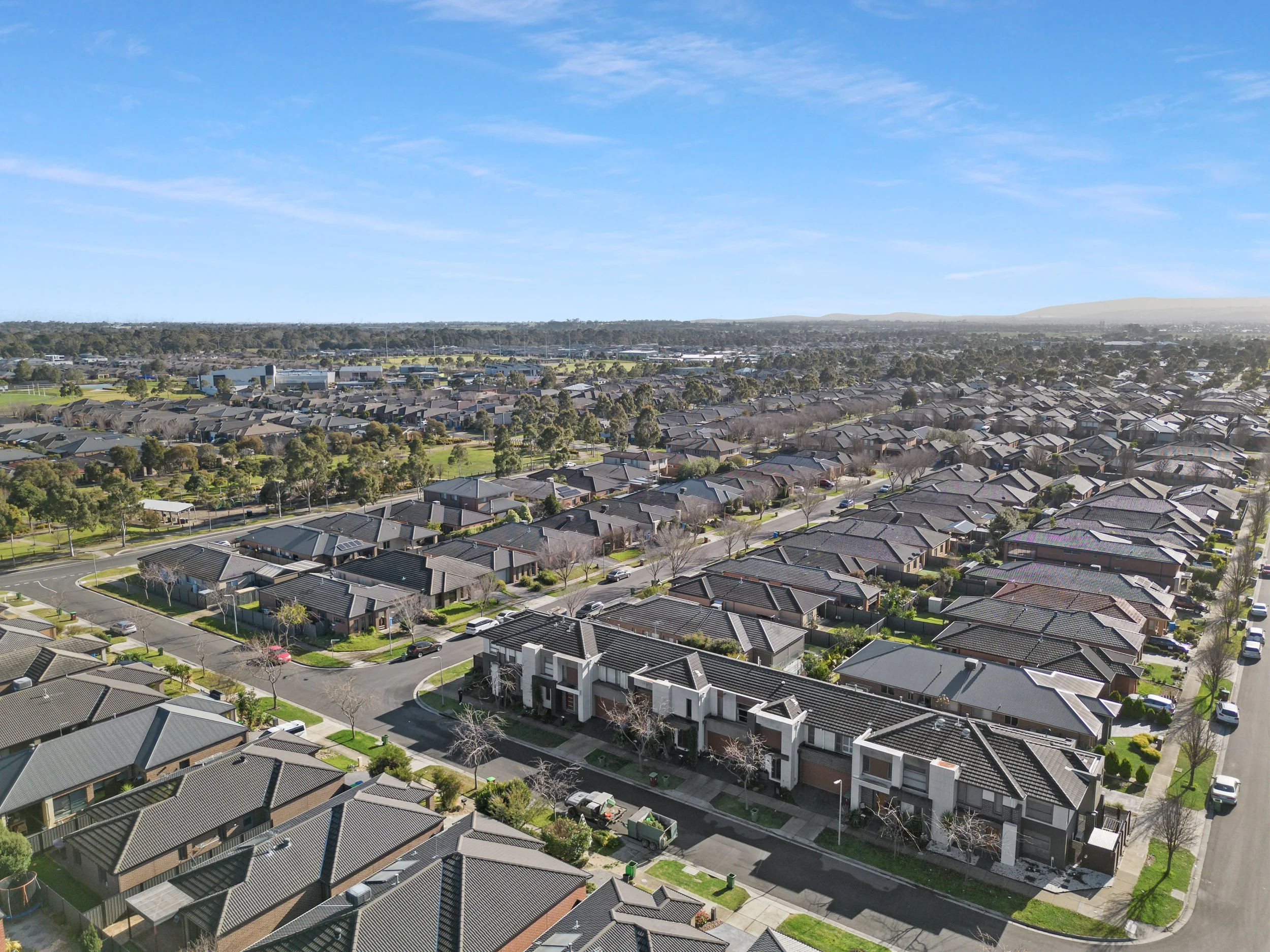 Aerial view of a suburban neighborhood with rows of houses, streets, and green spaces under a partly cloudy sky.